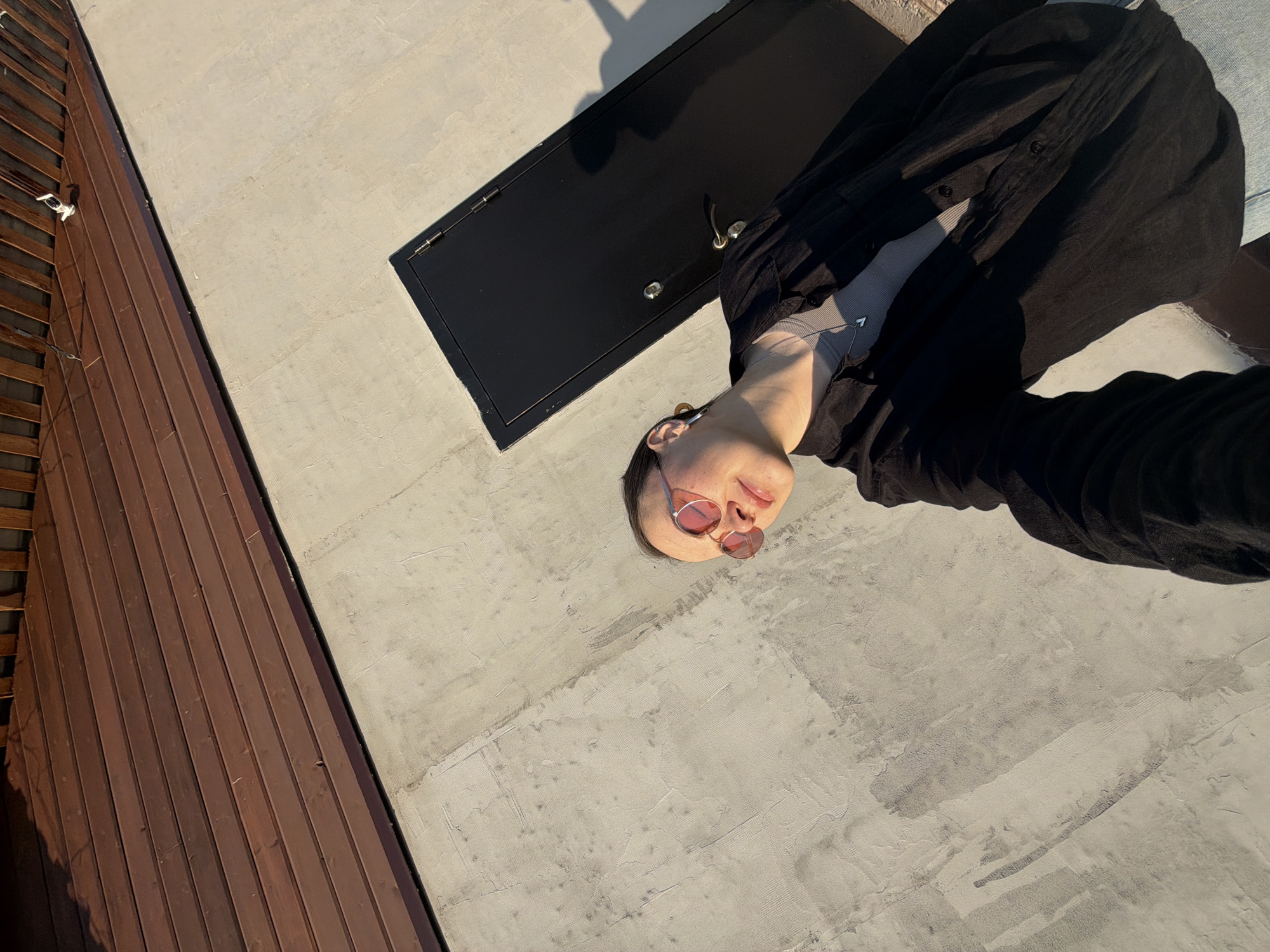 A young adult woman with fair skin and short brown hair stands on a concrete roof in Chaikovsky, Russia. She wears pink-tinted round sunglasses, a light-colored top, and a black button-down shirt. A black metal access panel lies adjacent to her. A wood-paneled structure is also visible on the roof. The scene is lit by natural sunlight.