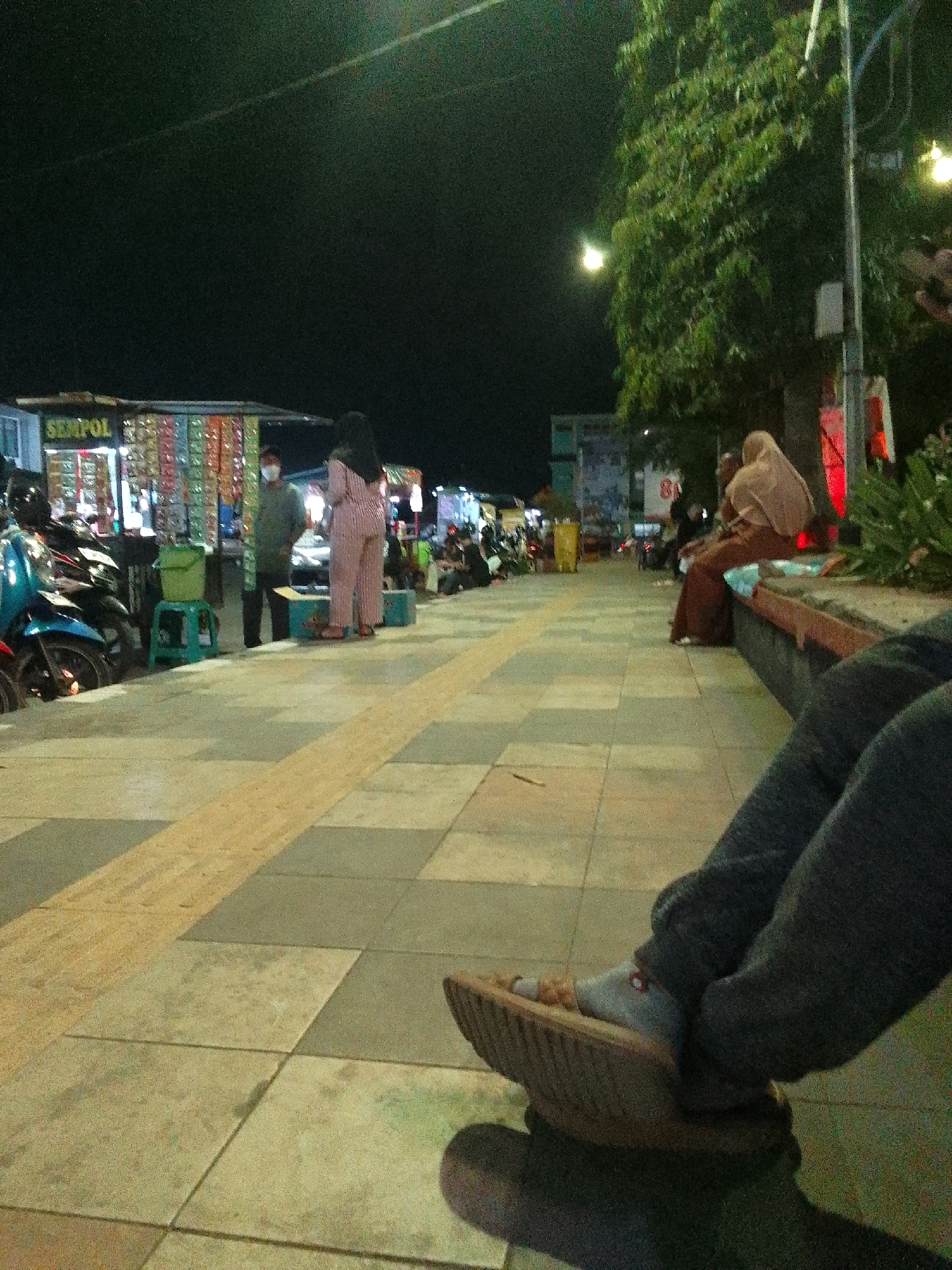 An outdoor, nighttime scene in Purbalingga, Indonesia, shows a pedestrian walkway with several individuals present. A stall with various items displayed is visible on the left, with motorcycles parked nearby. A person in a green shirt and a mask stands near the stall, next to a green bucket on a stool. Two women are also present; one in pink striped pajamas stands and another in a light brown headscarf and brown attire sits on a low wall to the right. In the foreground, a person's legs and feet are visible, with one foot wearing a sandal and a sock. The ground is paved with square tiles, with a yellow stripe running along the walkway. A large tree with dense foliage is on the right side of the frame, illuminated by streetlights.