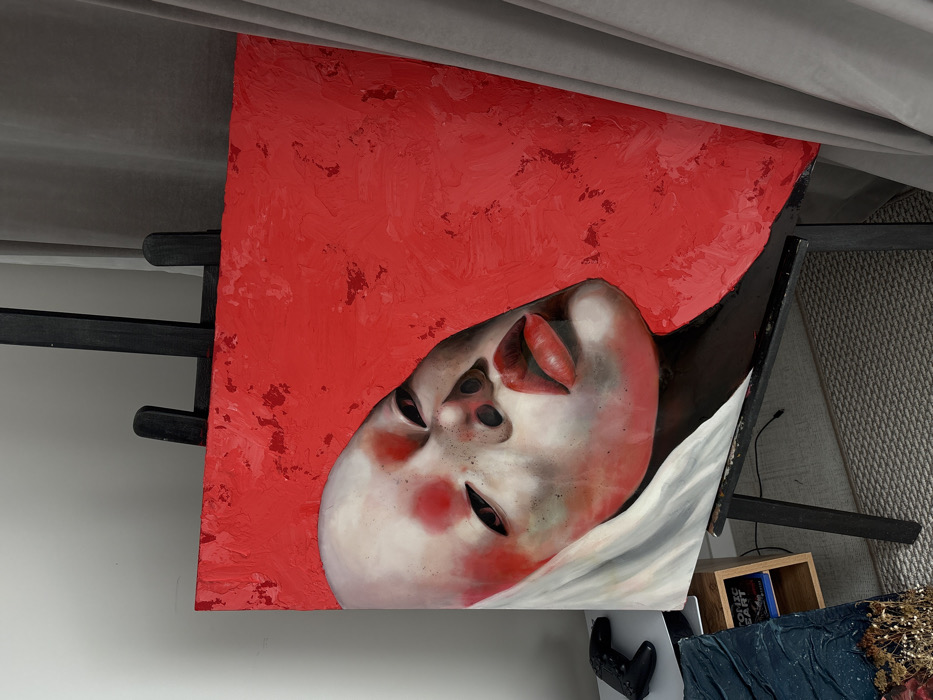 The image displays an indoor setting, likely a private residence or art studio, featuring a large, vertically oriented oil painting on a dark-colored easel positioned centrally. The painting depicts a stylized, pale human face or head, characterized by a smooth, possibly bald, cranial area. The facial features include dark, elongated eyes, prominent red lips, and areas of red pigmentation on the cheeks and forehead, with subtle dark smudging on the lower face and around the eyes. A white, draped element partially covers the left side of the subject's head. The background of the painting is a vibrant, textured red, rendered with visible impasto brushstrokes.
To the left of the easel, a black gaming controller rests on a light-colored surface. Below this surface, part of a light wooden shelf is visible, holding a book titled "ATOMIC ART" and another dark object. Further to the lower left, a portion of a second artwork is visible, exhibiting a dark blue, textured surface with embedded dried plant material. The floor is covered by a light grey patterned carpet or rug. On the right side of the image, light grey curtains hang, partially obscuring the background. The wall behind the controller and the upper left portion of the easel is a plain, pale grey.
There are no discernible architectural features, landmarks, or identifying elements within the image that specifically indicate its location within the city of Krasnoyarsk, Russia. The scene provides no information for geographical pinpointing beyond an interior space.