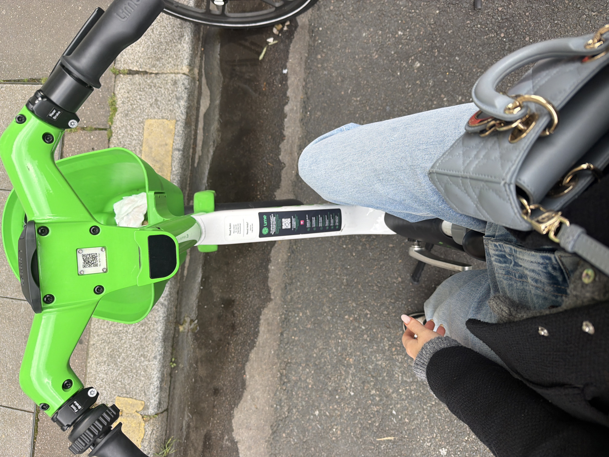 The image shows a top-down view of a person riding a Lime electric scooter. The scooter is predominantly white and green, with black handlebars and brakes. The person is wearing light blue jeans, a black coat, and a grey sweater underneath. A light grey quilted handbag with gold hardware is resting on the person's lap. The setting appears to be a street in a city, possibly Paris, France based on the scooter brand presence. The pavement is grey and wet, with a small section of yellow markings near the curb.