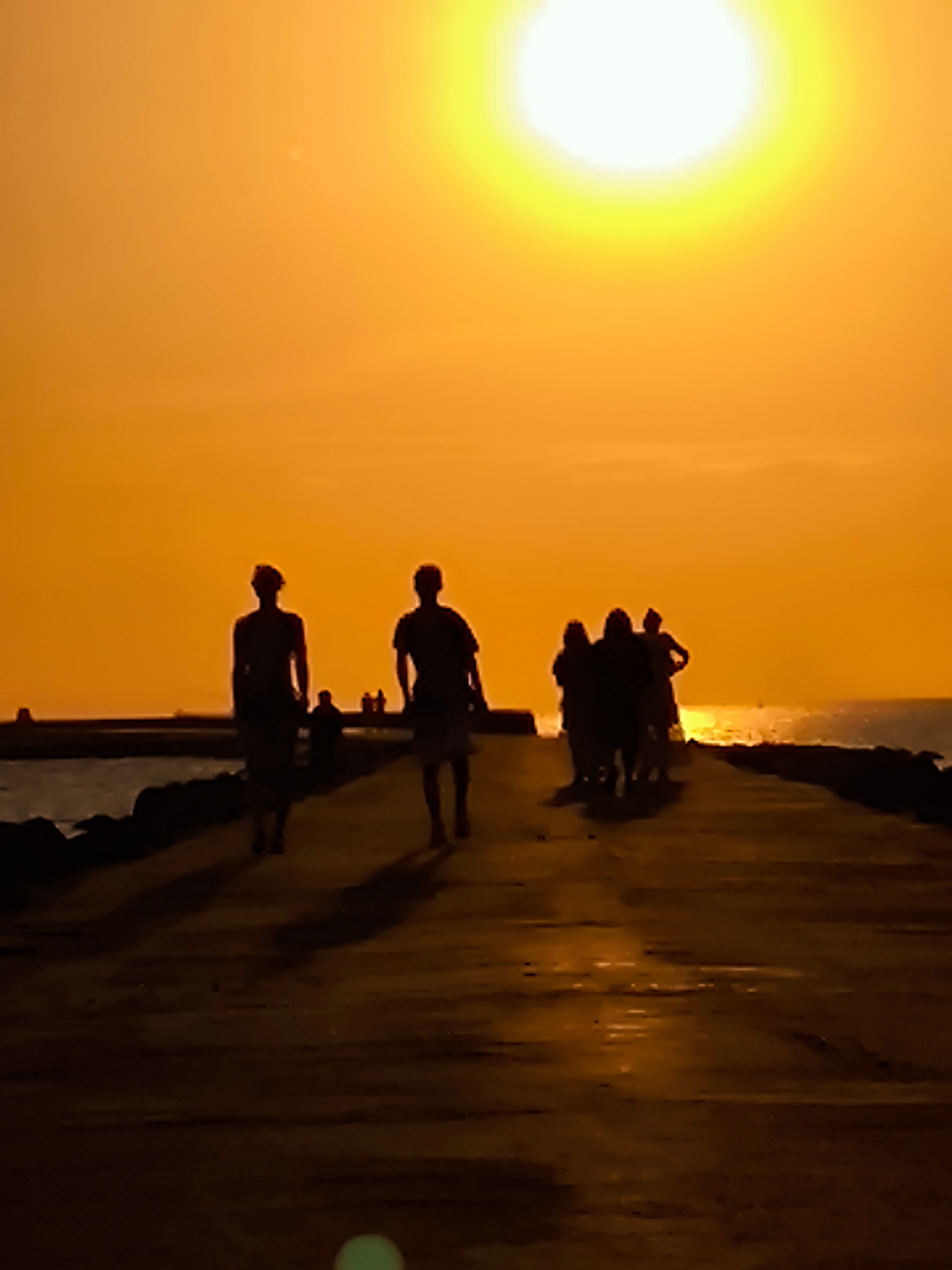A silhouetted group of people walk along a paved path next to a body of water, with a very bright sun setting in the background, casting an orange glow across the scene. The silhouettes are indistinct due to the backlighting. The path appears to be made of concrete or stone. The water on the left is dark and calm, with a rocky edge. The setting sun is a large, bright orb with a halo of light. The overall impression is of a warm, peaceful evening. This scene is not specifically located in Riga, Latvia.