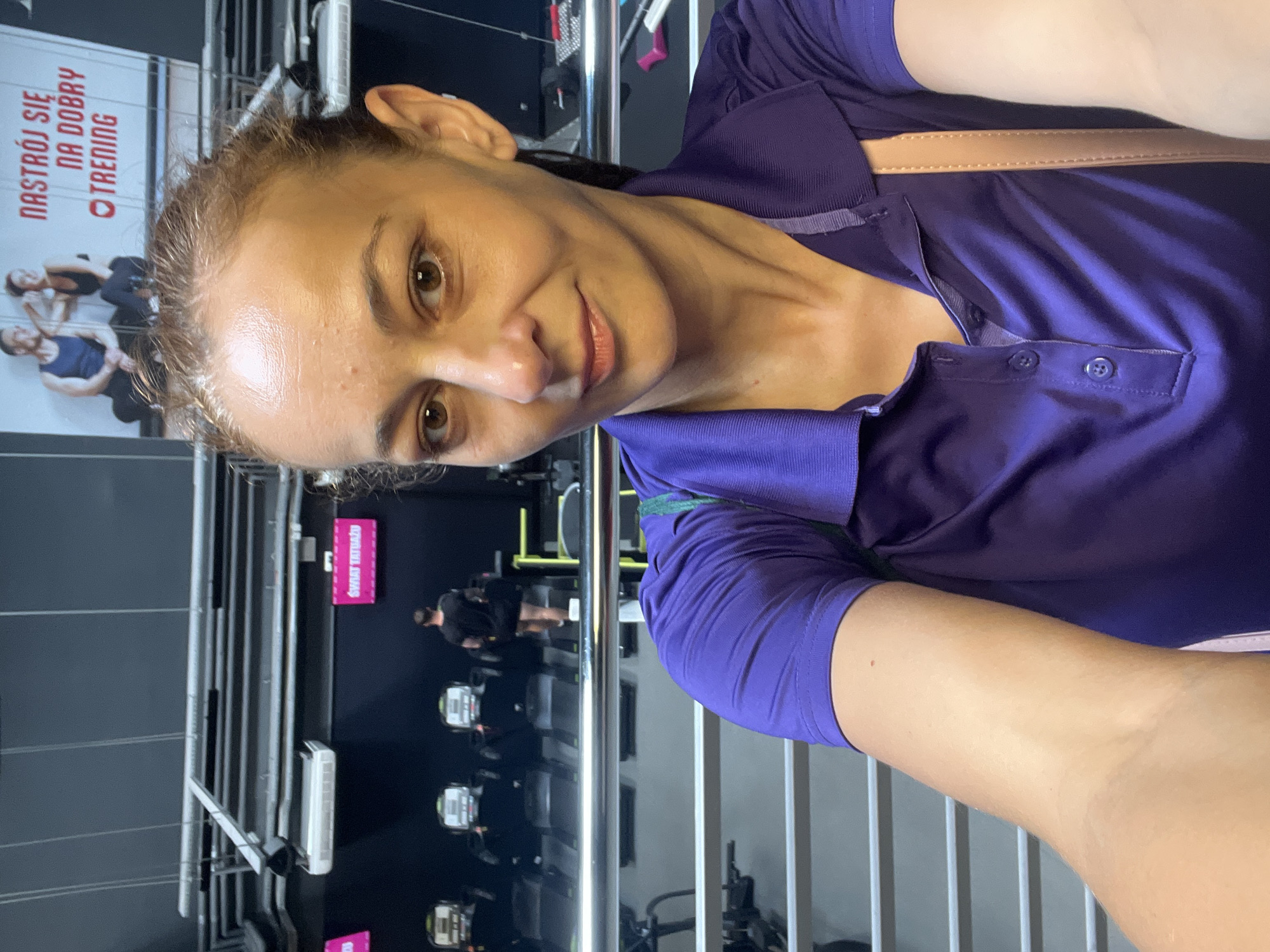A selfie is taken by a woman in a gym. The woman, who is wearing a purple polo shirt, is smiling and looking at the camera. In the background, there are several treadmills and a sign that says "NASTRÓJ SIĘ NA DOBRY TRENING," which translates to "GET IN THE MOOD FOR A GOOD WORKOUT." A man can be seen walking on one of the treadmills in the background. The gym appears to be well-lit and equipped with various exercise machines.