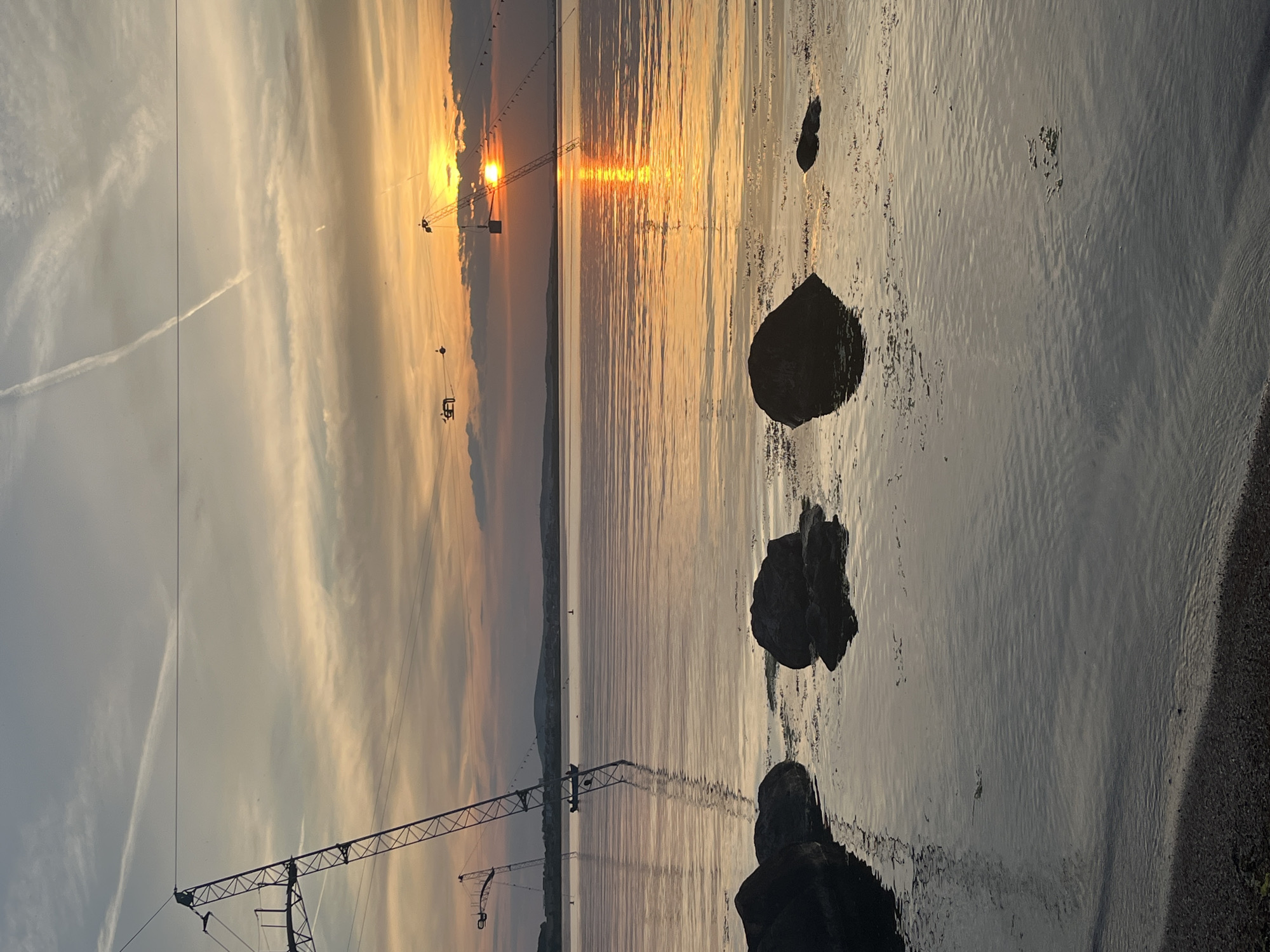 The image depicts a sunset over the sea, likely in the Burgas region of Bulgaria based on the presence of structures associated with water sports. The sky is filled with clouds, with the sun appearing as a bright orange orb near the horizon. Its reflection creates a shimmering path on the water's surface. Several large rocks are visible along the shoreline in the foreground, partially submerged in the water. A metal structure, possibly a wakeboarding or waterskiing cable system, stretches along the water. A portion of a crane is visible.