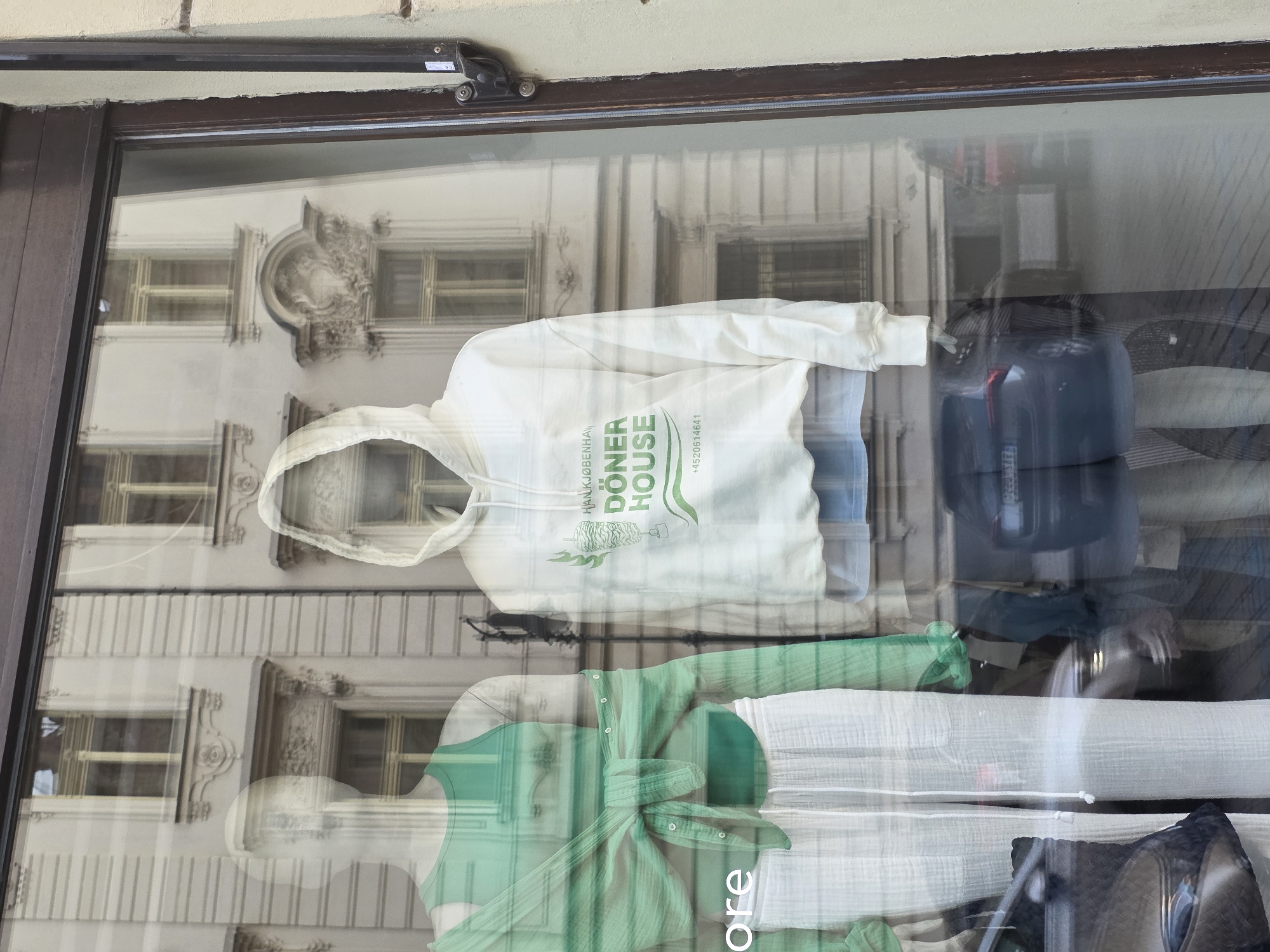 The image is a medium shot of a storefront window display. Two mannequins are visible. The mannequin on the left is wearing a green tank top layered with a sheer green open-button shirt tied at the waist, and white gauze pants. The mannequin on the right is wearing a white hooded sweatshirt with green text and graphics that read "HANKJØBENHA DÖNER HOUSE" and a phone number "+4520614641". The background of the display is a reflection of an ornate building with windows. Outside the window, cars and a street are visible, suggesting an urban setting. The overall lighting is neutral, with reflections from the building and street obscuring some details.