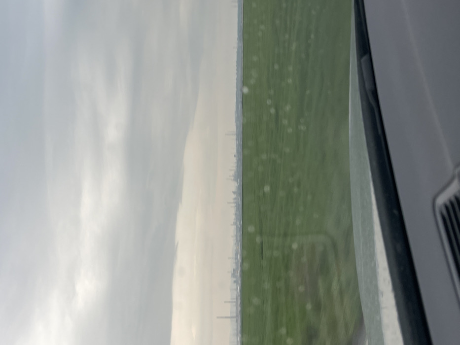 The image displays a wide, exterior view from the interior of a vehicle, specifically over its dashboard and through a windshield. The bottom approximately 15% of the frame shows a dark gray dashboard and the base of a windshield. The windshield exhibits numerous small, circular, bright specks, consistent with raindrops.
The mid-foreground, spanning approximately 40% of the image, is dominated by an expansive, level green field, appearing to be cultivated vegetation. This field extends horizontally across the frame.
Above the green field, stretching across the horizon line, is a distant industrial complex located in Bratovo, Bulgaria. This complex is characterized by a dense array of numerous tall, slender smokestacks, towers, and other industrial structures, appearing somewhat blurred and hazy due to distance and atmospheric conditions.
The upper approximately 30% of the frame consists of an overcast sky, presenting a uniform light gray hue with minimal cloud definition, suggesting a dull or cloudy day. No individuals or discernible actions are present in the scene.