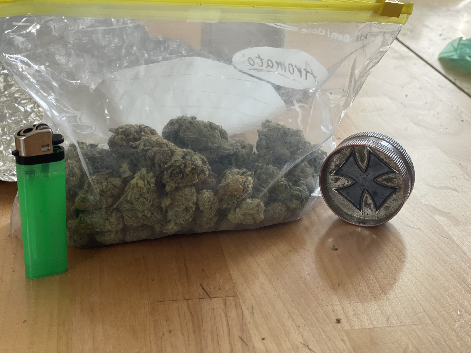 A green lighter, a clear plastic bag containing cannabis buds and a white substance, and a metal grinder with a star emblem are arranged on a wooden surface. The plastic bag is sealed with a yellow zipper closure. Text is visible on the bag, appearing to be handwritten in black marker. A crumpled piece of foil is visible behind the plastic bag. The lighting suggests the scene is indoors, possibly on a table or floor.