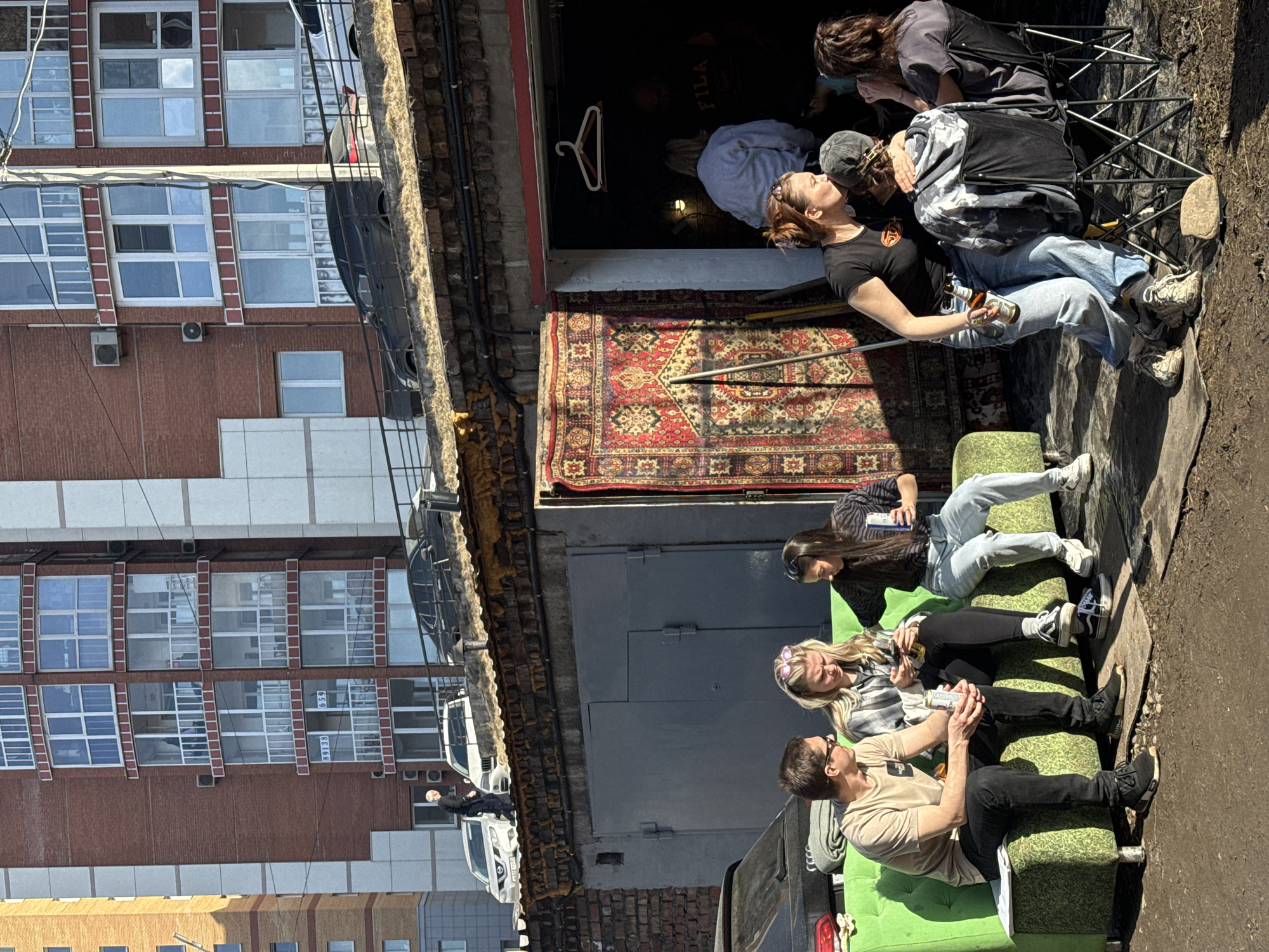 The image depicts a group of people gathered outdoors, appearing to be socializing and relaxing in an urban setting. Three people are seated on a green couch. Two other individuals are seated behind on folding camp chairs, holding bottles of alcohol. One holds a beer, and another is embracing this person. The setting is adjacent to a building, with a decorative rug hung at the entryway. The building's structure includes brickwork and concrete elements, typical of urban architecture. Outside the building is a parking lot of vehicles. Buildings are located across the street. The scene suggests a casual, informal gathering in an urban environment. Visible building numbering along the facade indicates the location in Krasnoyarsk, Russia, is likely the corner of Ulitsa Uritskogo and Prospekt Mira.