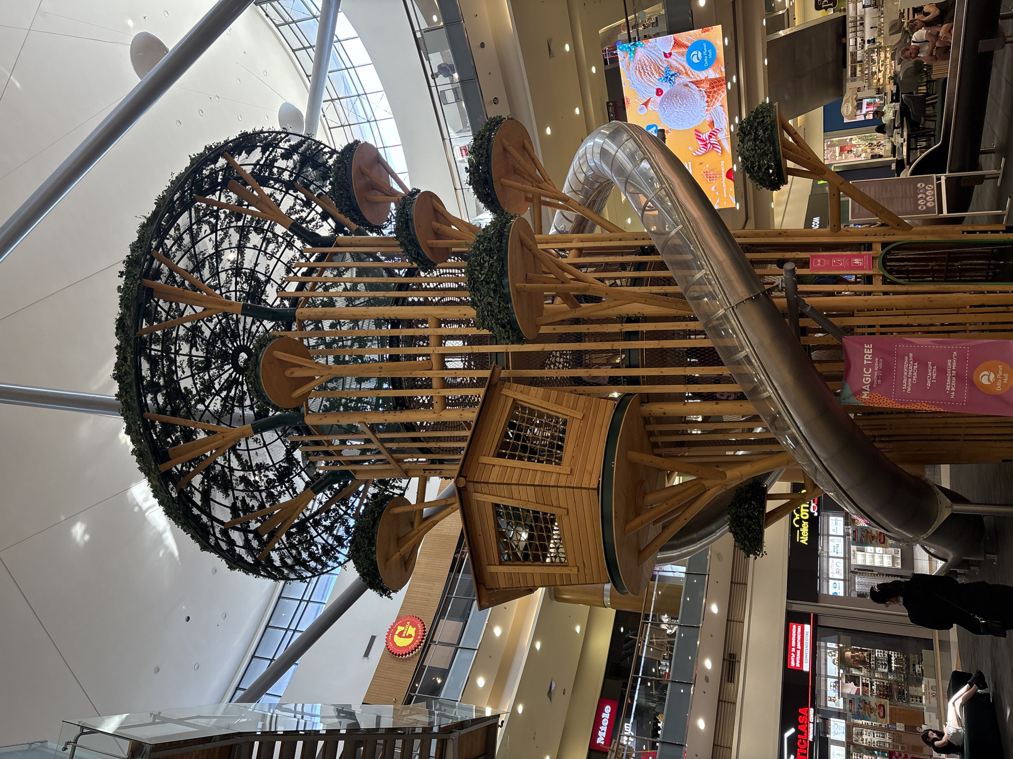 This image shows a large, whimsical wooden playground structure inside a shopping mall. The structure, designed to resemble a "Magic Tree," features a large net-like sphere covered in greenery, multiple wooden platforms with decorative trees, and a winding metal slide. A small wooden house with windows is nestled within the structure. The mall environment is visible in the background with shops and escalators. Two people are partially visible at the bottom right of the image. The lighting suggests it is daytime. Several signs in Bulgarian are visible, including one that says "MAGIC TREE" and provides rules for entry, such as mandatory personal protective equipment, a 2-meter distance, and disinfection every 30 minutes. Other signs include shop names like "Miele" and "Atelier OTI." The overall scene is that of a family-friendly entertainment area within a commercial space.