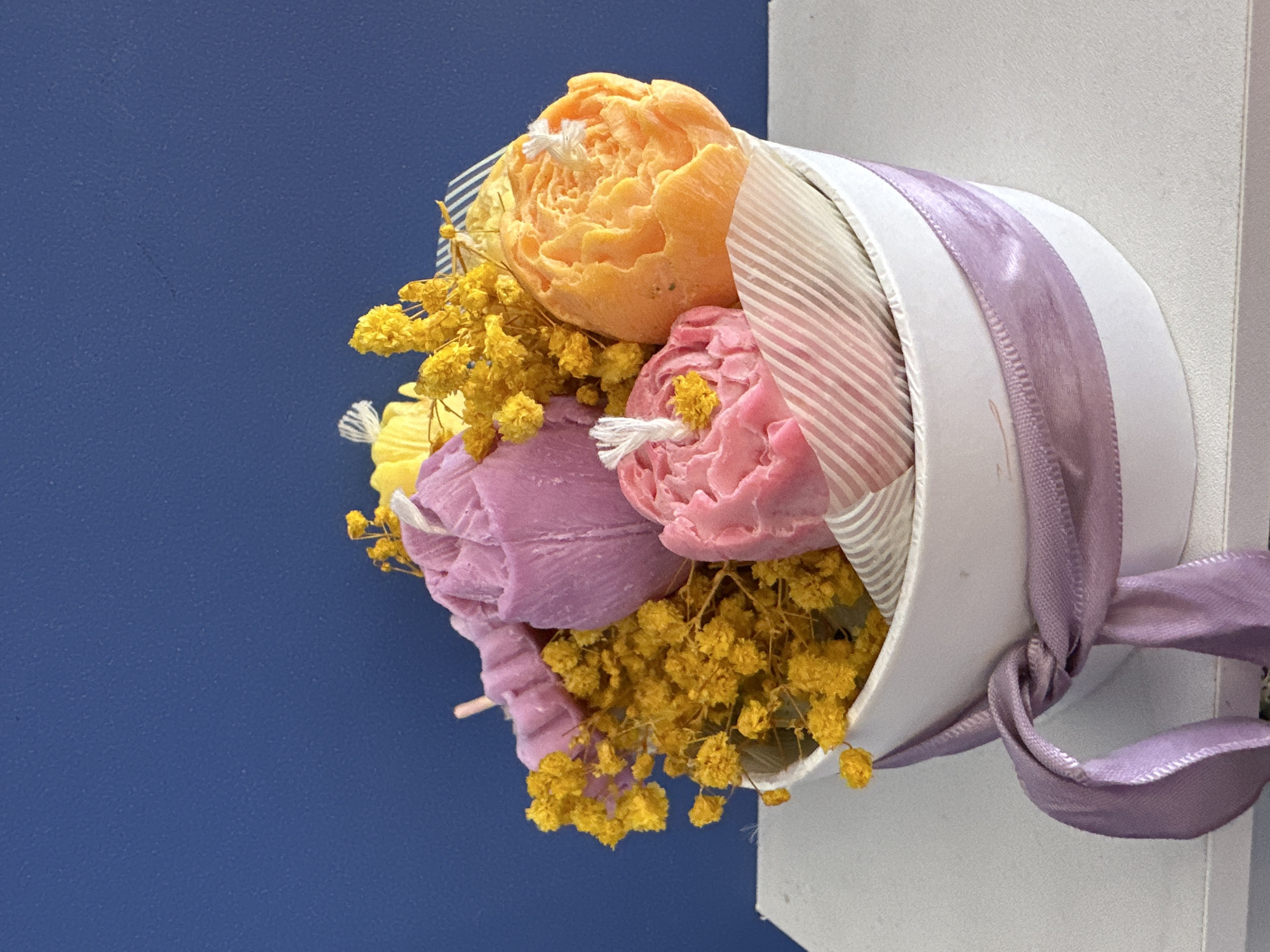 A still life shows a floral arrangement in a white container, tied with a lavender ribbon, against a split background of dark blue on the left and white on the right. The floral arrangement features three faux flower candles, one orange, one pink, and one purple, interspersed with yellow dried flowers. The candle wicks are white. The white container has layers of striped decorative paper inside. The lavender ribbon is tied in a bow. The blue background is plain, and the white background appears to be a wall. The image is shot at an angle.