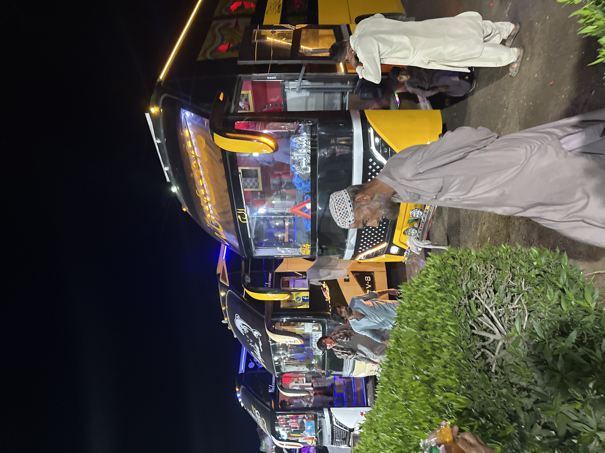 The image captures a nighttime scene at what appears to be a bus terminal or stop. Several men are present, gathered around multiple large passenger buses.
The main subjects are the people and the buses. An older man with a beard, wearing a light grey shalwar kameez and a white and red prayer cap, stands prominently in the foreground, looking towards the buses. Other men, also dressed in traditional attire, are visible, some standing near the buses, and one is seen bending down.
The scene is dominated by at least three brightly lit buses, primarily black with distinctive yellow accents. They appear to be modern, possibly double-decker, with illuminated interiors and external decorative lights, including blue and yellow LED strips. The activities suggest passengers waiting, boarding, or disembarking. One person is visible in the bottom right, holding a small bottle or container near a dense green bush.
Notable details include the clear nighttime setting, indicated by the dark sky and the glow from the buses and surrounding artificial lights. The clothing worn by the men suggests a South Asian or Middle Eastern cultural context. Visible text on the buses includes "V-B", "Welcome" on the side windows and near the front windshields, and several lines of Arabic script. One prominent piece of Arabic text appears to be "الرحم" (Al-Rahim, potentially part of Al-Rahman). The overall mood is calm, reflecting routine travel activities.