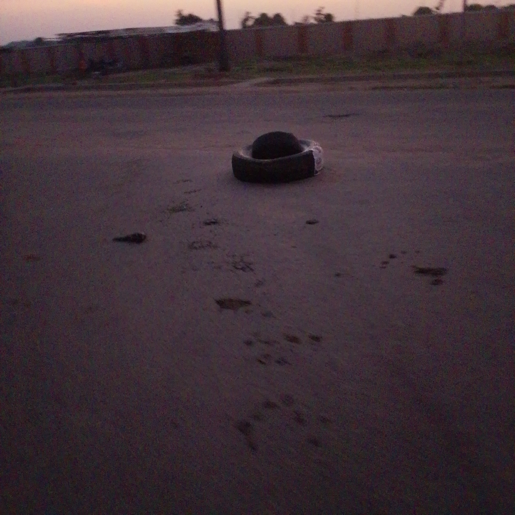 A tire lies on a dark, paved surface, possibly a road, in Wuro Allahamdu, Nigeria. The sky is a dusky, pale orange, suggesting dawn or dusk. In the background, a low, dark structure with a fence can be seen, with some vegetation and trees behind it. The ground in the foreground shows some indistinct marks and patches, possibly footprints or debris, and a small, dark object is visible to the left of the tire. The overall scene is dimly lit and somewhat blurry.