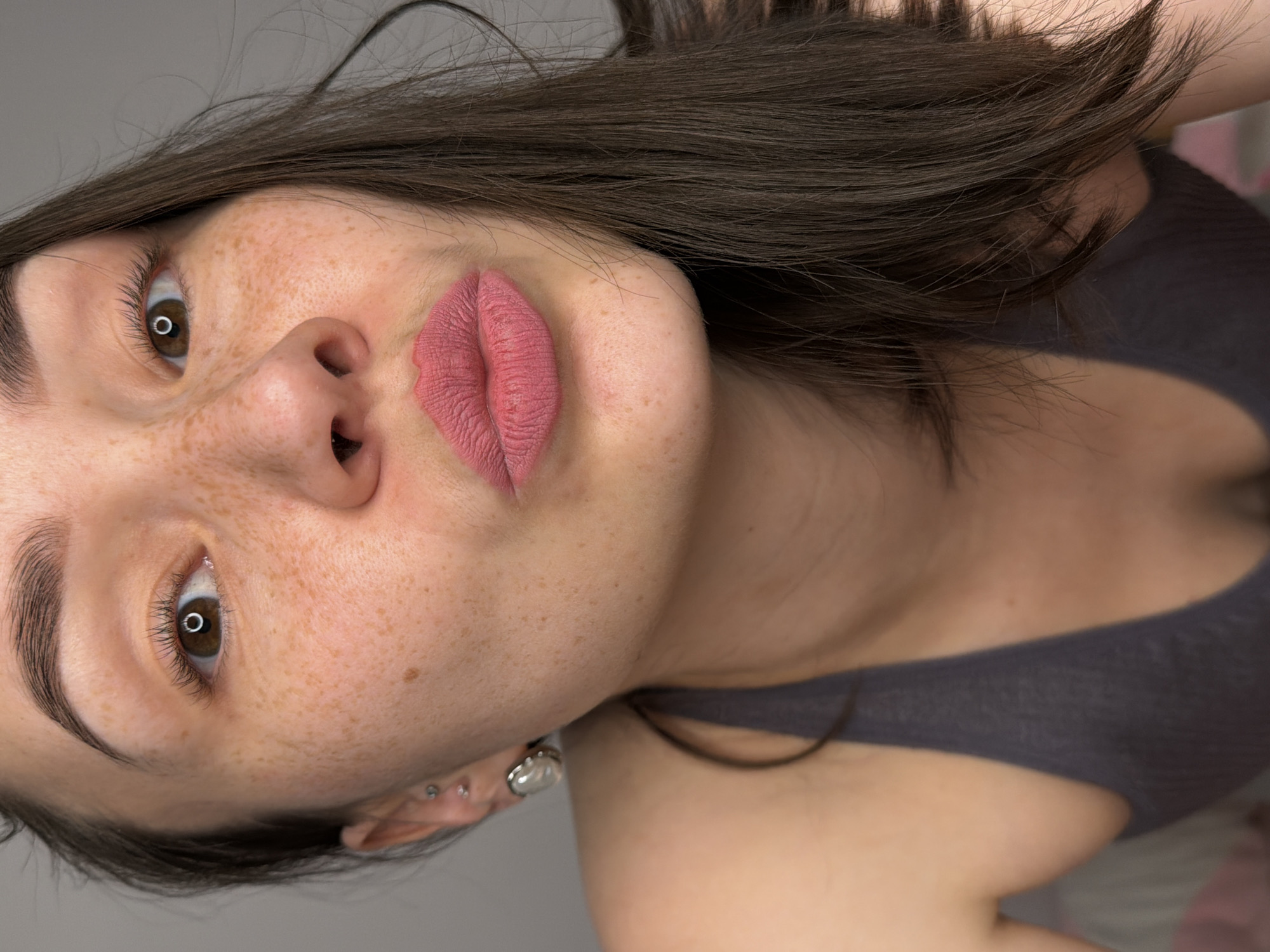 A close-up, slightly angled portrait captures a young woman with light skin, brown hair, and freckles. She has brown eyes and is wearing a muted pink lipstick. Her hair is styled loosely around her face. The subject is wearing a gray top. The background is a plain, neutral color. The image also shows a pierced ear, including jewelry worn.
This image does not contain enough information to determine if it was taken in the city of Chaikovsky, Russia. The setting is minimal and indistinct.
