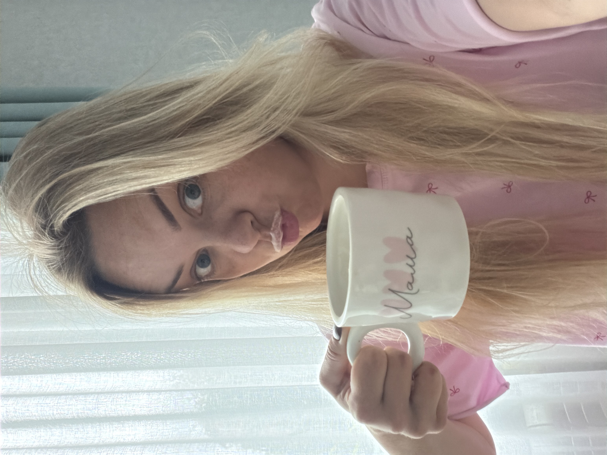 A blonde woman with milk around her upper lip holds a white mug with "Mama" written on it and pink hearts above it. She is wearing a pink t-shirt with small pink bows on it. The woman is looking at the camera with a surprised expression and her eyes are wide open. The background is a window with white vertical blinds. The image appears to be taken indoors in a home environment, possibly Votkinsk, Russia, though no specific location details are evident in the image itself.