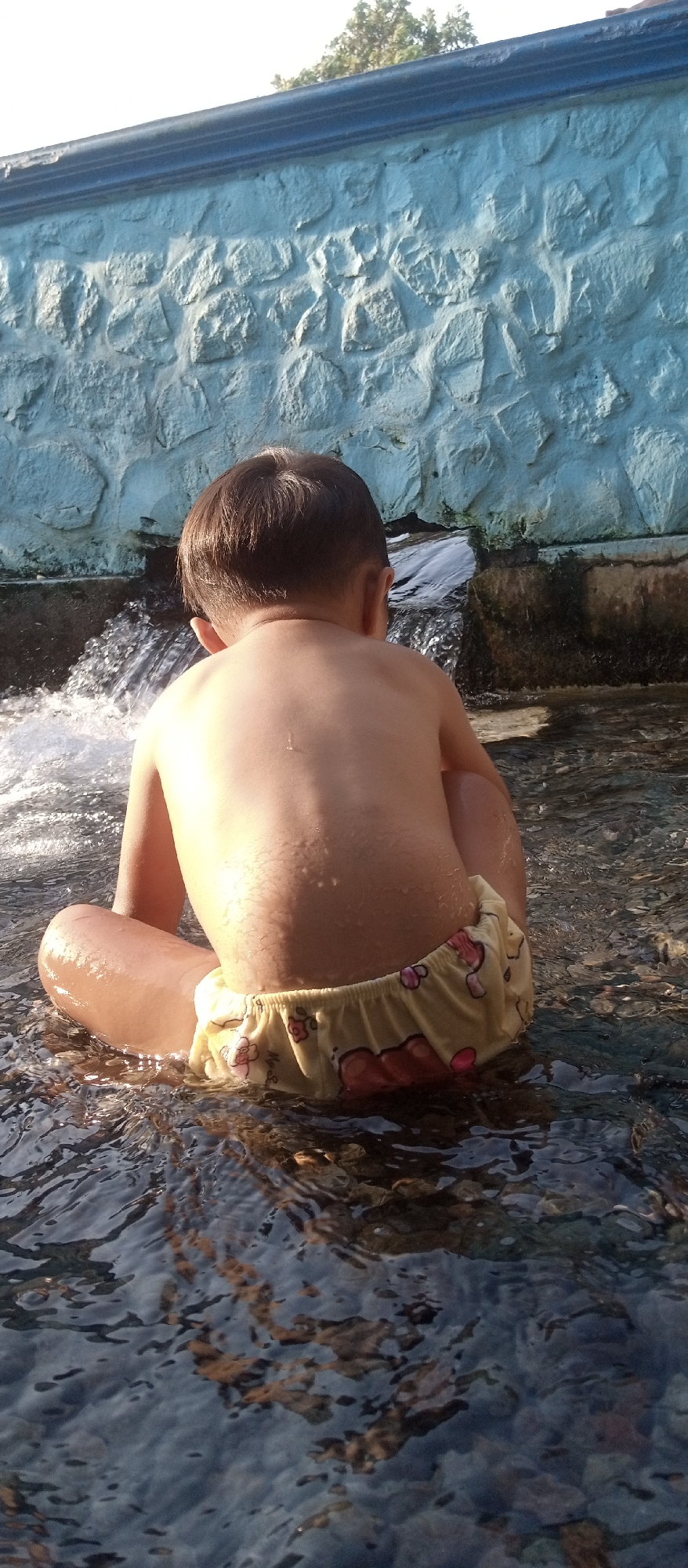 A young child is sitting in shallow water, facing away from the camera. The child is wearing light-colored shorts with a floral pattern. The water is flowing gently, and the child appears to be playing or cooling off. Behind the child is a textured, light blue wall, possibly made of stone or concrete. The environment suggests an outdoor setting, perhaps a public park or a residential area in Udanwuh, Indonesia, given the specific details of the wall and the typical warm climate that would encourage such activity. The sunlight creates reflections on the water's surface.