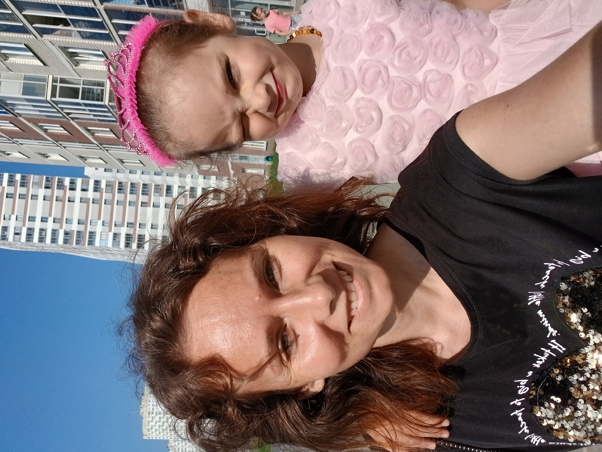 In a selfie taken outdoors, two females are captured with modern apartment buildings and a bright blue sky in the background, suggesting an urban environment in Yekaterinburg, Russia. The female in the foreground, on the left, is an adult with wavy brown hair, smiling broadly with her eyes open. She is wearing a black t-shirt adorned with sparkling sequins arranged in a heart shape and white script lettering. Her left arm is visible, resting on or near the shoulder of the other individual.
To her right and slightly behind her is a young girl, likely a child, sporting a pink tiara with sparkly accents. She is wearing a pale pink, frilly dress with rosette-like detailing. The girl is winking and smiling, with some glitter on her cheek. She is also wearing a necklace made of brown beads.
In the background, behind the girl, a third person, a child or young teenager, is visible from the waist up, wearing a pink top and denim shorts, facing away from the camera. The architectural style of the buildings suggests a newer development area. The lighting indicates it is daytime, likely sunny.