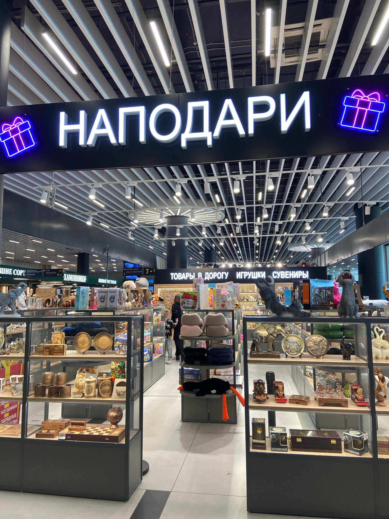The image shows the exterior of a gift shop in Ob, Russia, with the shop's name, "НАПОДАРИ" (NAPODARI), displayed in large white illuminated letters above the entrance. The sign is flanked by neon outlines of gift boxes. Inside, various items are displayed in glass cabinets, including chess sets, decorative plates, figurines of elephants and other animals, and wooden boxes. Several people, including children and adults, are visible browsing the merchandise. The shop appears to be in an indoor shopping area, indicated by the ceiling with exposed pipes and lighting fixtures, and the presence of other storefronts in the background.