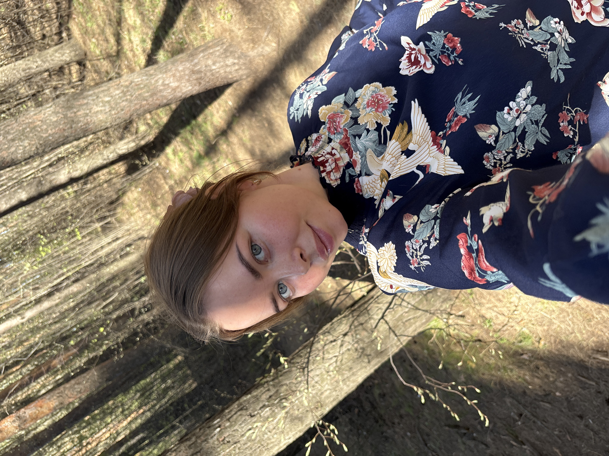 A young woman is the primary subject of the image, appearing to be taking a selfie outdoors. She has fair skin, light brown hair, and light-colored eyes. She is wearing a dark blue blouse with a floral and crane print. The background consists of tree trunks and sparse vegetation, suggesting a wooded area, likely within the city of Izhevsk, Russia. The lighting indicates it is daytime, possibly early spring given the budding trees. The angle of the photograph is from a slightly upward perspective, typical of a selfie.