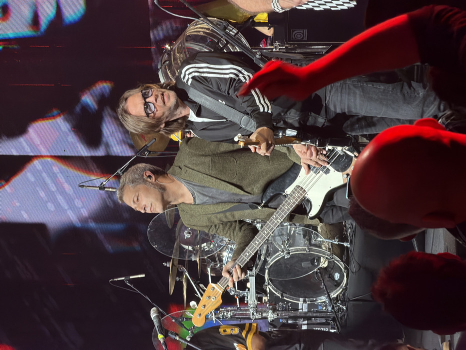 This image captures a live musical performance at a concert venue in Tel-Aviv, Israel. The specific venue is not identifiable from the visual evidence.
In the foreground, the heads of several audience members are visible, illuminated by a strong red stage light. One audience member's arm is raised vertically, also glowing red.
The central focus is on two male musicians on stage. The musician on the left, with short graying hair and an in-ear monitor in his right ear, wears an olive-green blazer over a dark grey t-shirt. He holds a light-colored four-string bass guitar with a white pickguard, his left hand on the fretboard and his right hand near the bridge. He is looking upwards and to the left. The musician on the right has medium-length brown hair and wears dark sunglasses with blue lenses, a black track jacket with three white stripes on the sleeves, and dark jeans. He holds a dark electric guitar, his left hand on the neck and his right hand near the body. He is looking forward and slightly to the right, with his mouth slightly open.
Behind the two main musicians, elements of a drum kit are visible, including multiple drums, cymbals, and clear acrylic sound shields. Several microphones on stands are positioned across the stage. To the far right, an amplifier cabinet with a circular speaker grille is visible. The stage floor is covered with various cables.
The background consists of a large LED screen or projection displaying abstract, colorful patterns with vertical and horizontal lines, and a stylized, distorted image of a face or figure in hues of red, blue, and white. The stage is brightly illuminated by overhead lighting, casting reflections on the instruments and equipment.
