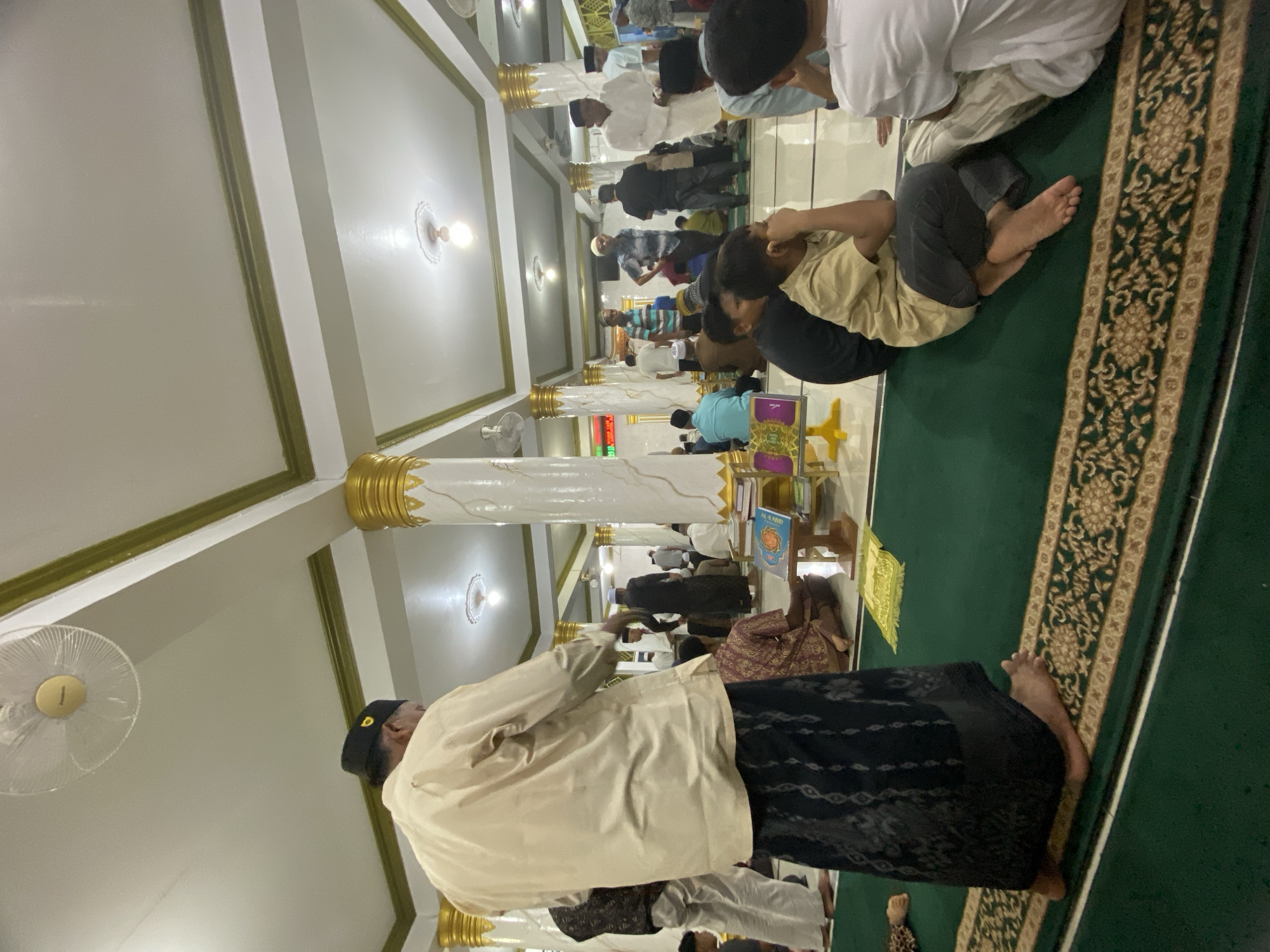 The image captures an interior view of a mosque in Lhokseumawe, Indonesia, likely during a gathering or prayer session. In the foreground, a man wearing a light-colored shirt and a dark patterned sarong stands facing away from the camera. Several other individuals, predominantly men and boys, are seated on a green carpet adorned with a decorative border. Some are engaged in prayer or contemplation, while others appear to be conversing. The mosque's architecture is visible, featuring white walls, high ceilings with ornate gold accents on the columns, and lighting fixtures. In the background, more people are present, and some architectural elements of the mosque are discernible. The overall atmosphere suggests a communal religious setting.