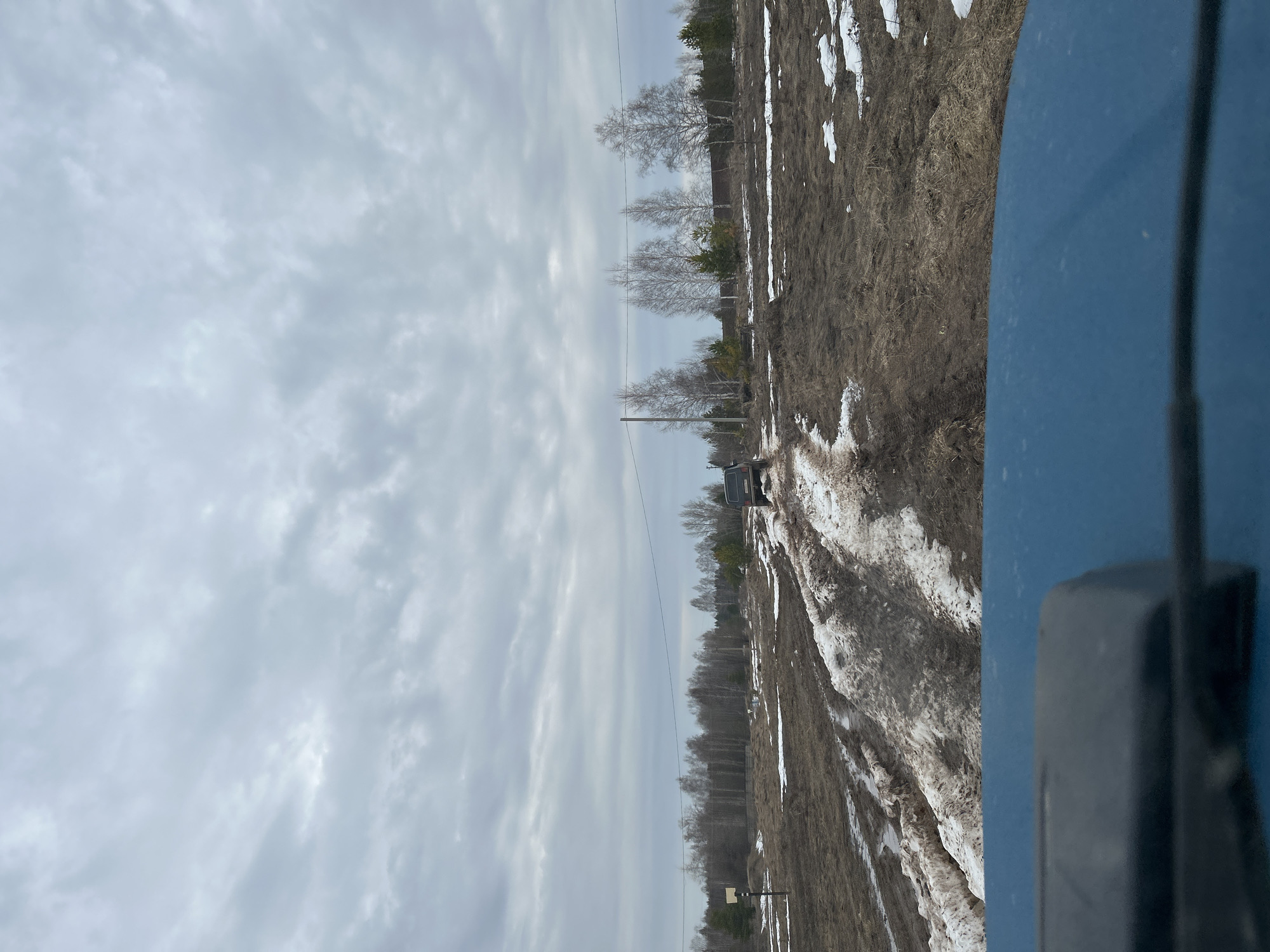 The image shows a rural dirt road landscape under an overcast sky. Patches of melting snow are visible along the roadside and in the ruts of the road. The right side of the frame includes part of a blue vehicle, with a wiper arm visible. A dark-colored vehicle is further down the road. Power lines stretch across the scene. Bare trees line the horizon. The location is most likely Уральское, Russia.