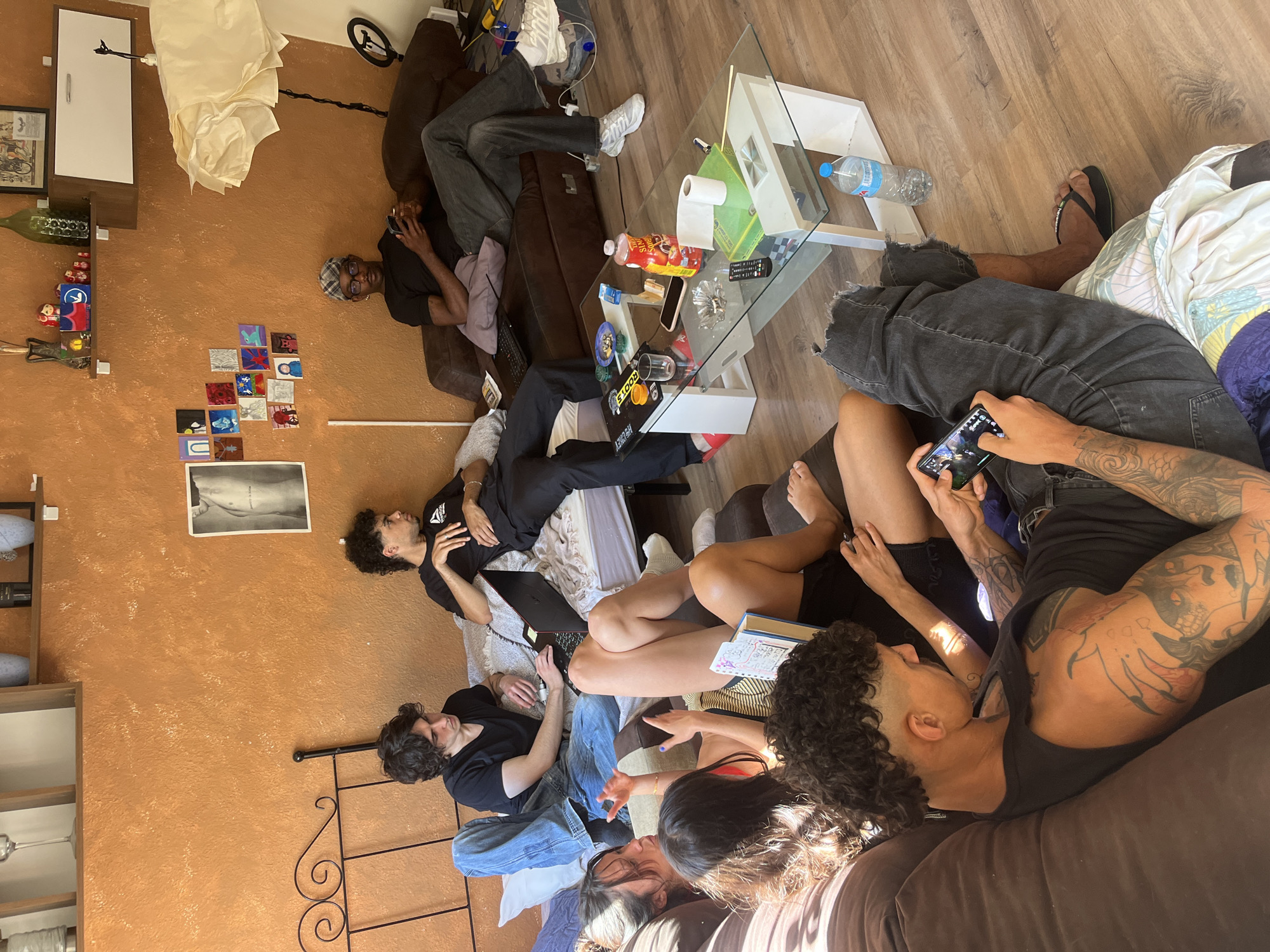 The image depicts a group of people casually lounging indoors. There are at least eight individuals visible, some on couches, others on what appears to be a bed or futon. They are engaged in various activities, including using phones, laptops, and talking among themselves. A glass-topped coffee table is in the center, with items like a water bottle, snacks, and remote controls on it. The room has warm-toned walls, and a shelf with decorative items is mounted on one wall. There's also a piece of artwork hanging on the wall. Based on the style of furniture, architecture, and presence of individuals with European features, the location could plausibly be Barcelona, Spain, but there are no specific details to make a definitive determination.