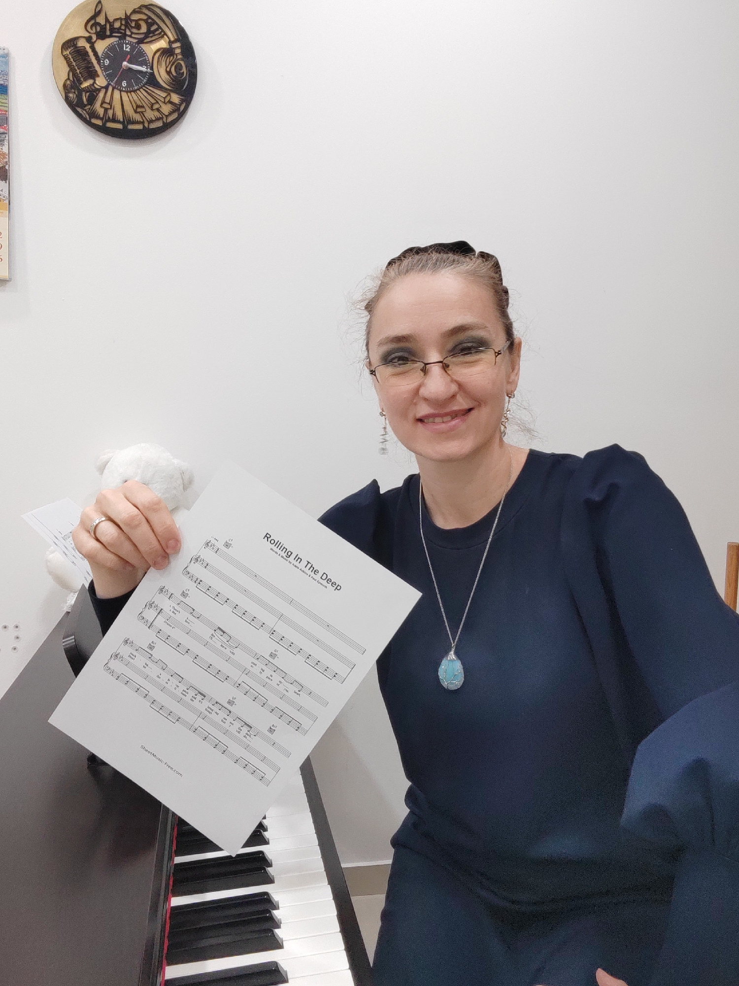A smiling woman, appearing to be in her 40s or 50s, is captured in a close-up portrait, seated at a digital piano or keyboard in an indoor setting in Podgorica, Montenegro. She wears glasses, a dark long-sleeved top, and a distinctive necklace with a light blue/turquoise pendant. Her hair is pulled back, and she has dark eye makeup, contributing to her warm, inviting expression.
The woman holds up a sheet of music prominently towards the viewer, displaying the title "Rolling In The Deep" by Adele Adkins and Paul Epworth. The music sheet also features a "SheetMusic-Free.com" watermark at the bottom. Behind the sheet music, a small white plush toy, possibly a teddy bear, is partially visible.
The scene includes the black and white keys of a digital piano or keyboard in the foreground and to her right. On the white wall behind her, a unique round wall clock with a musical theme is mounted, depicting a microphone, headphones, piano keys, and musical notes. The clock's hands indicate a time, though precisely readable. A sliver of a calendar or another framed item with numbers (2, 6, 5) is visible on the far left wall. The lighting is bright and even, typical of an indoor daytime setting. The woman's joyful smile suggests enthusiasm or welcome.