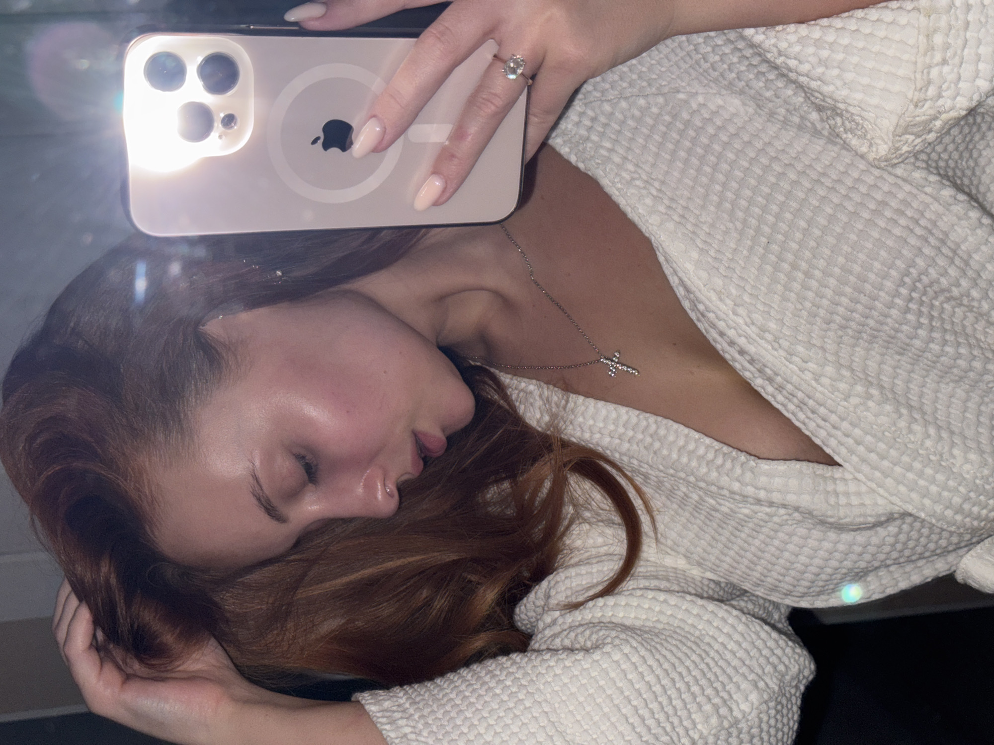 A medium-shot portrait depicts a woman with reddish-brown hair and fair skin, lying on a surface. She is wearing a white waffle-patterned robe and a silver necklace with a cross pendant. The woman's eyes are closed, and her head is resting on her hand. Above her head, a hand holds an iPhone with the camera facing downwards, causing a bright reflection. A diamond ring is visible on the finger holding the phone. The location of the image is unknown, as is its connection to the city of Chaikovsky, Russia.