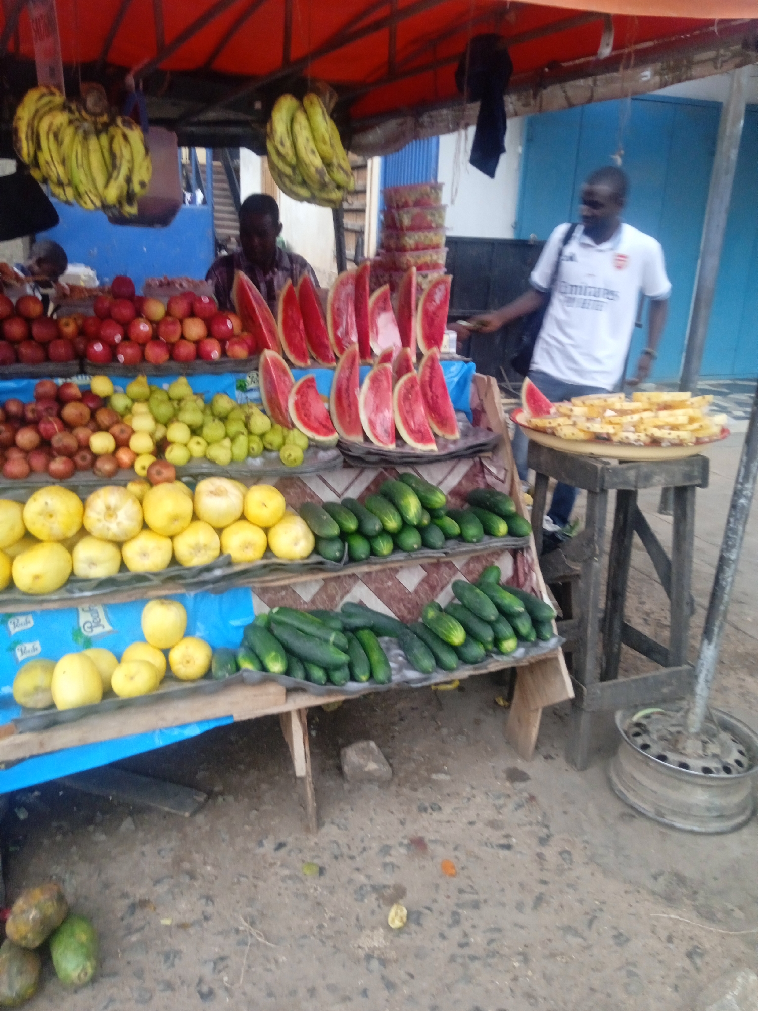 A bustling fruit and vegetable stand dominates the scene, likely located in Jos, Nigeria. The stand displays a colorful assortment of produce, including hanging bunches of bananas, rows of red apples, green pears, yellow lemons, vertically arranged watermelon slices, and piles of green cucumbers. A second stand, set up behind it, is showing sliced pineapple. The vendor stands behind the main display. A customer, wearing a white t-shirt, stands nearby, possibly making a purchase. The setting is likely an outdoor market or roadside stand, given the makeshift nature of the display and the outdoor environment.