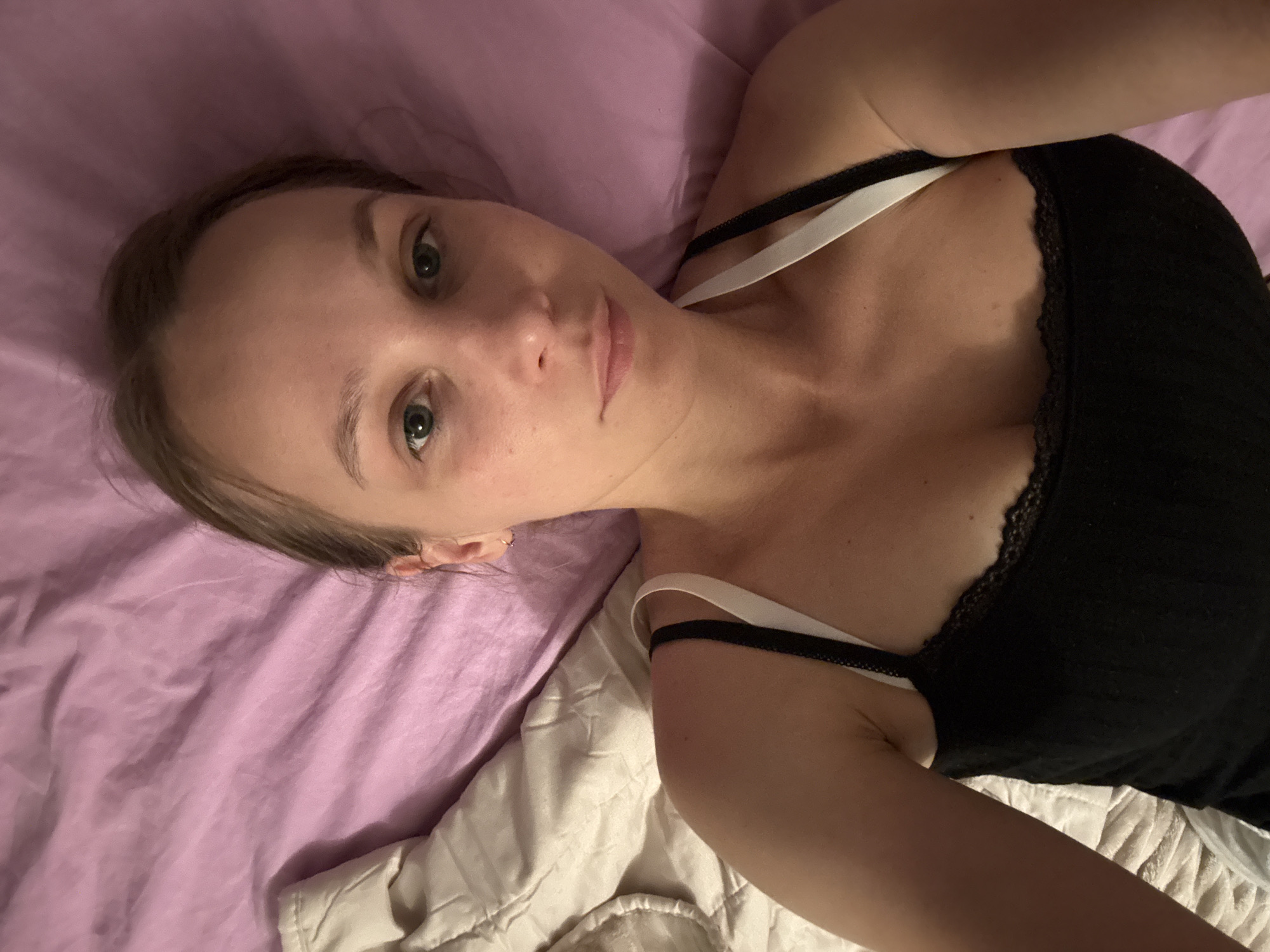 A young woman with light skin and green eyes lies on a bed with lilac-colored sheets and a white duvet. She is wearing a black camisole with lace trim. Her head is tilted back, and she is looking directly at the camera. Her hair is light brown and pulled back from her face. The lighting suggests an indoor setting, possibly during the evening or night, as there is a soft glow to the scene. The overall mood appears relaxed and intimate. There is no visible text in the image. The location context is Überherrn, Germany.