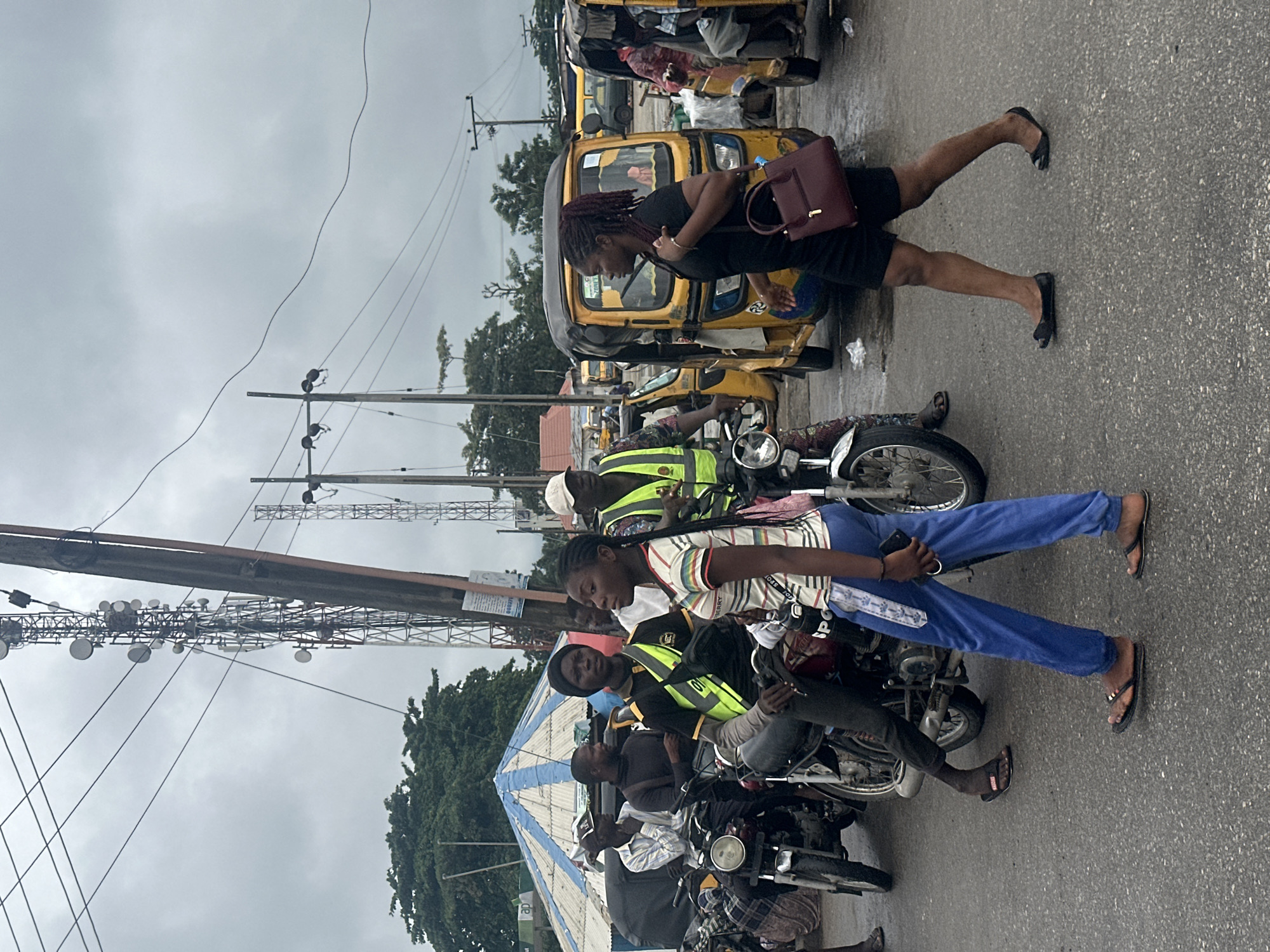 The image depicts a street scene in Badagry, Nigeria, under a cloudy, overcast sky. The primary subjects are several people, some of whom are interacting with yellow auto-rickshaws, commonly known as "keke." In the foreground, a young woman wearing a striped t-shirt and blue pants is seated on a motorcycle, holding a phone. Beside her, a man in a black shirt and reflective yellow vest is also on a motorcycle.
Further back, another woman with red braids, wearing a black t-shirt and shorts, stands next to a keke, holding a burgundy handbag. She appears to be either about to enter or exit the vehicle. Several other kekes are parked or moving in the background, suggesting a transportation hub or a busy street. The presence of power lines and poles indicates an urban or semi-urban environment. The overall atmosphere seems to be one of daily activity and movement. The time of day is not explicitly clear due to the cloud cover, but it appears to be daylight. No specific text is legible in the image.