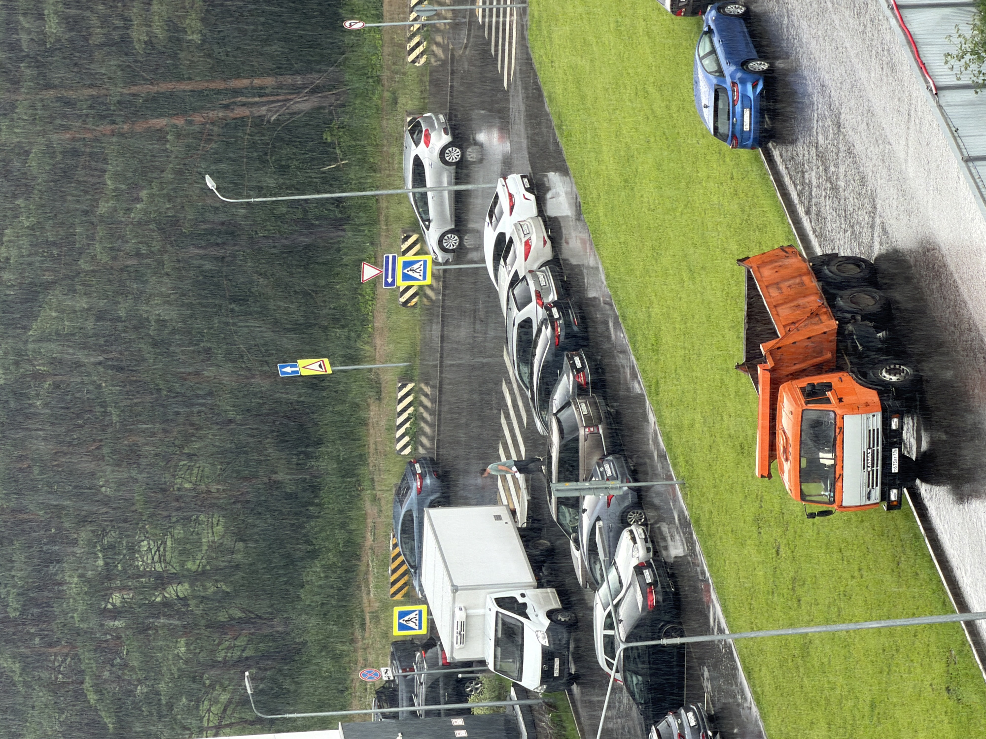 A high-angle, eye-level shot captures a rainy day in an urban environment. In the foreground, an orange dump truck is parked on a wet asphalt surface. To the left of the truck, a neatly mown green lawn borders the asphalt. A line of cars, including a blue hatchback, a silver SUV, a dark sedan, and a white sedan, are parked along the side of the road. In the middle ground, a white box truck is parked on a paved area, with a person standing near its rear. Several other cars are parked behind the box truck. A crosswalk is visible on the wet pavement. Road signs, including a pedestrian crossing sign and a warning sign, are present. In the background, a dense line of trees forms a natural barrier. The sky is overcast, and heavy rain is falling, indicated by visible streaks across the image and the wet surfaces. Streetlights are positioned throughout the scene.