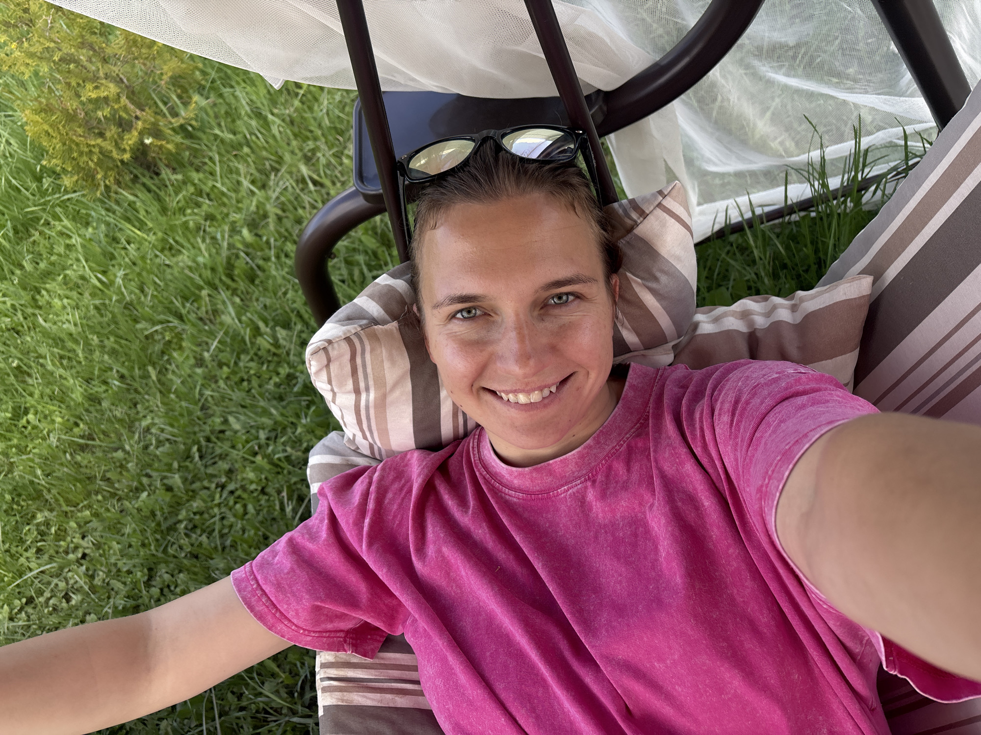 A person is lying on a striped cushion with a pillow under their head in what appears to be a swing or hammock. They are wearing a pink t-shirt and have sunglasses perched on their head. The person is smiling at the camera. The background includes green grass and foliage, and the frame of the swing or hammock with a light-colored canopy. This photo was taken outdoors in Раздолье, Russia.