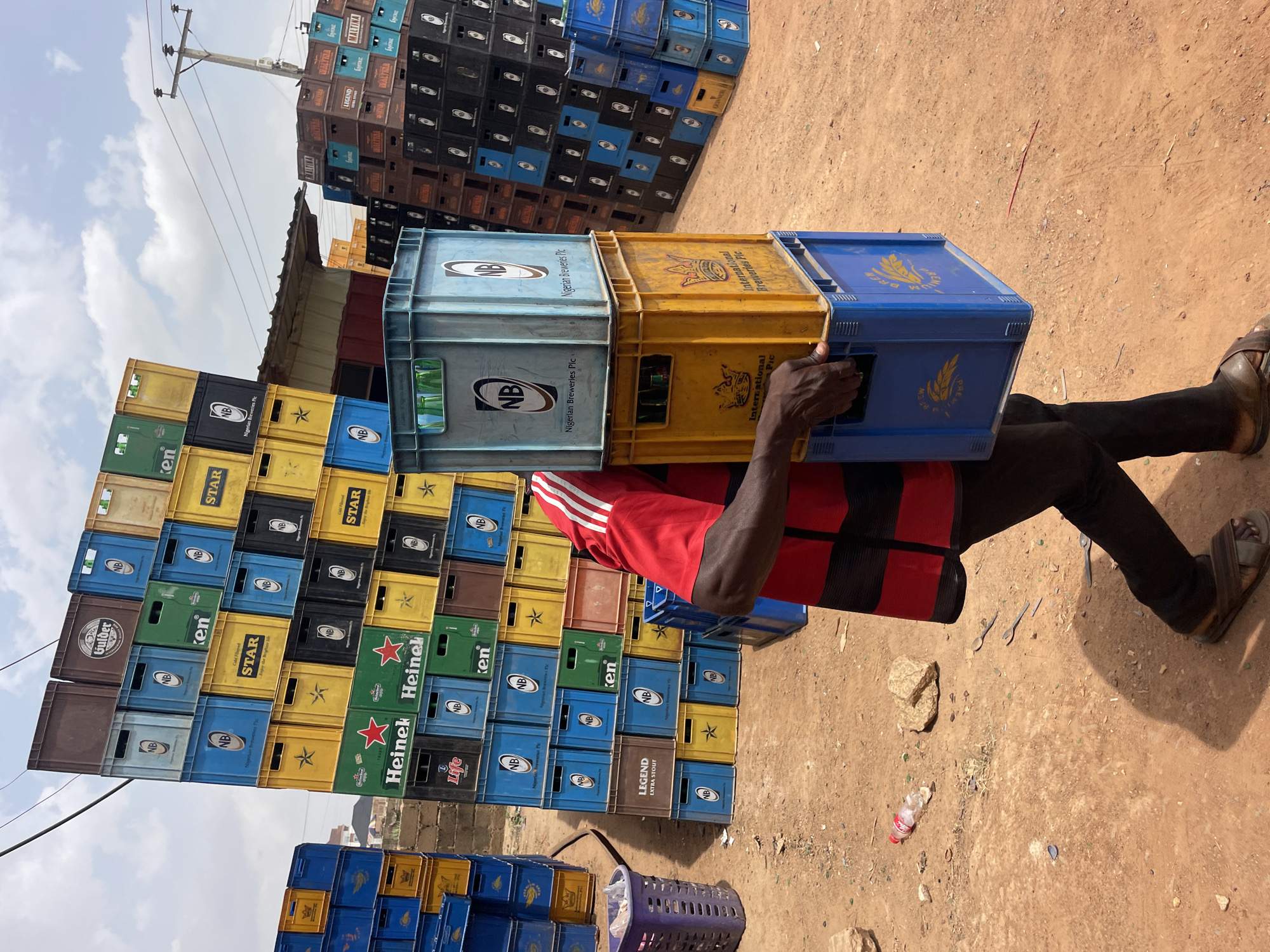 The image depicts a man in Gura Top, Nigeria, transporting a stack of three beer crates on his head. He wears black trousers, brown sandals, and a red shirt with white stripes. He is walking on a dirt ground littered with a plastic bottle, small rocks, and discarded metal spoons.
Behind him are large stacks of beer crates of various colors and brands, including NB (Nigerian Breweries), Star, Heineken, Legend, Gulder, and Ken. A small, makeshift shelter is partially visible behind the stacks. In the distance are buildings and a utility pole with power lines against a partly cloudy sky.