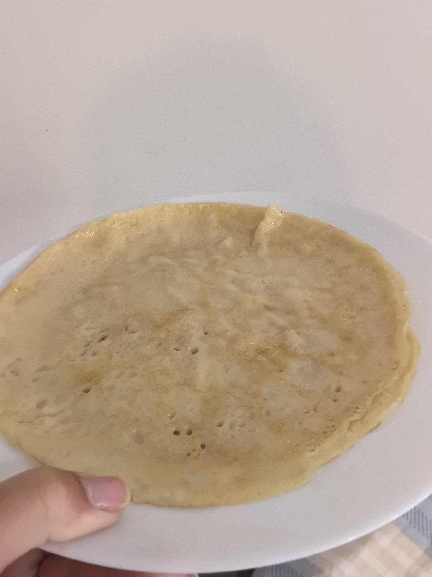 A close-up image captures a single, round pancake or crepe resting on a white plate. The food item is a light golden-brown color, with a slightly uneven surface featuring numerous small indentations and subtle variations in texture, suggesting it has been freshly cooked. Its edges are slightly irregular.
A human hand, visible in the lower-left corner, gently supports the edge of the white, rimmed plate. The visible nails on the hand are neatly manicured and painted a glossy light pink.
The background behind the plate and food is a plain, light off-white surface, likely a wall, creating a simple and uncluttered setting. In the bottom right corner of the frame, a small section of a plaid or checkered tablecloth, featuring light blue and white or beige squares, is visible, indicating the plate is placed on a table. The lighting in the image is soft and even, consistent with indoor conditions. There is no visible text or explicit indication of time of day.