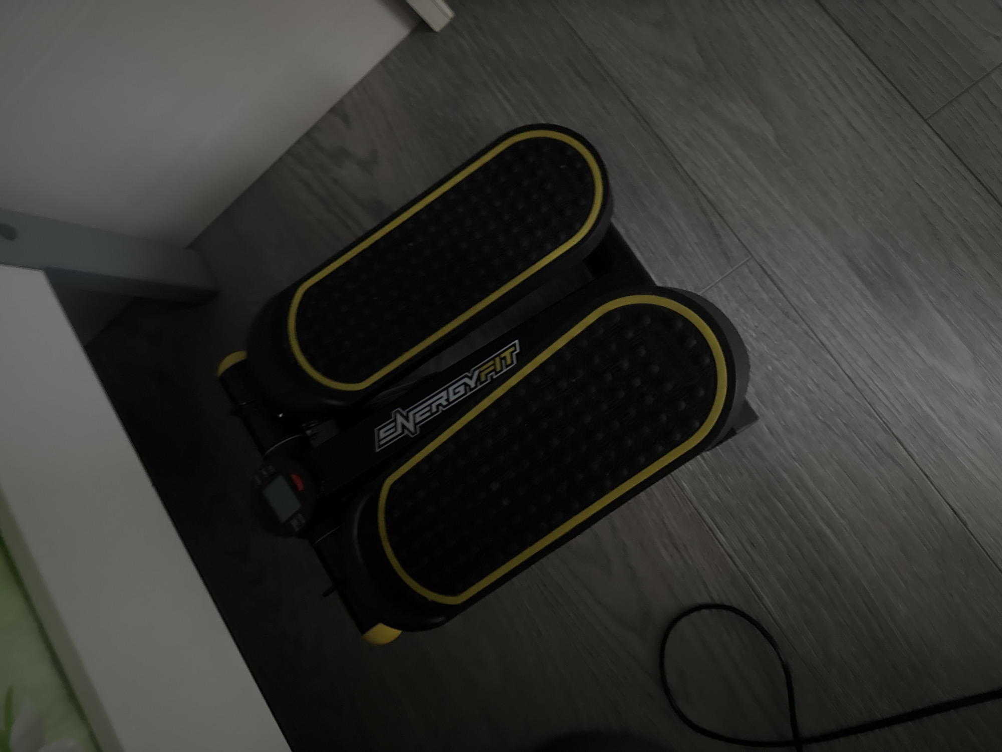 A black and yellow mini stepper machine is positioned on a gray laminate floor. The stepper has two footplates with textured surfaces and yellow trim. A small digital display is located at the front of the machine. A black cord loops from the left side of the stepper onto the floor. The background shows the base of a white piece of furniture and a gray vertical support. The overall setting appears to be indoors, possibly a home. The image does not provide any information to infer its location in Mikhaylovsk, Russia.