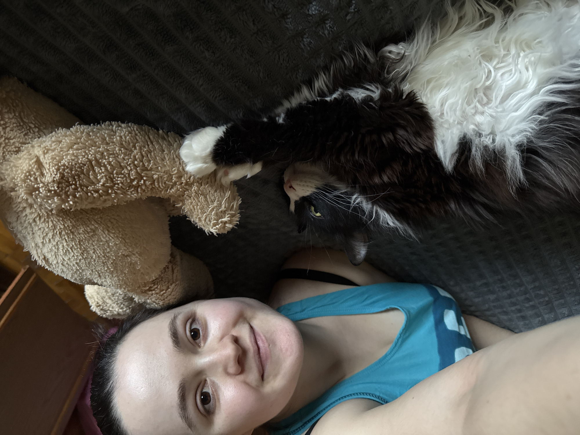 A woman is taking a selfie with a long-haired black and white cat and a brown teddy bear on a gray textured blanket. The woman is wearing a blue tank top. The cat is lying upside down with one paw touching the teddy bear. The specific location in Izhevsk, Russia, is not identifiable from the image.