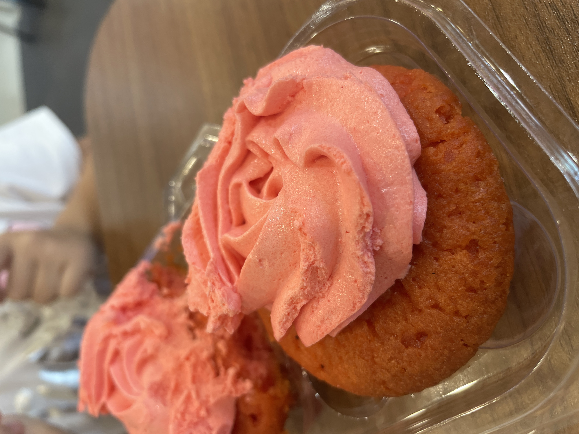 A close-up shot displays two cupcakes with pink frosting, housed within a clear plastic container. The cupcake on the right is fully assembled with a swirl of pink frosting resembling a rose, while the cupcake on the left has its frosting partially removed. The cupcakes appear to be sitting on a wooden table indoors. A person’s hands are slightly visible in the background, out of focus. The location in the city of Nha Trang, Vietnam cannot be determined from the image.