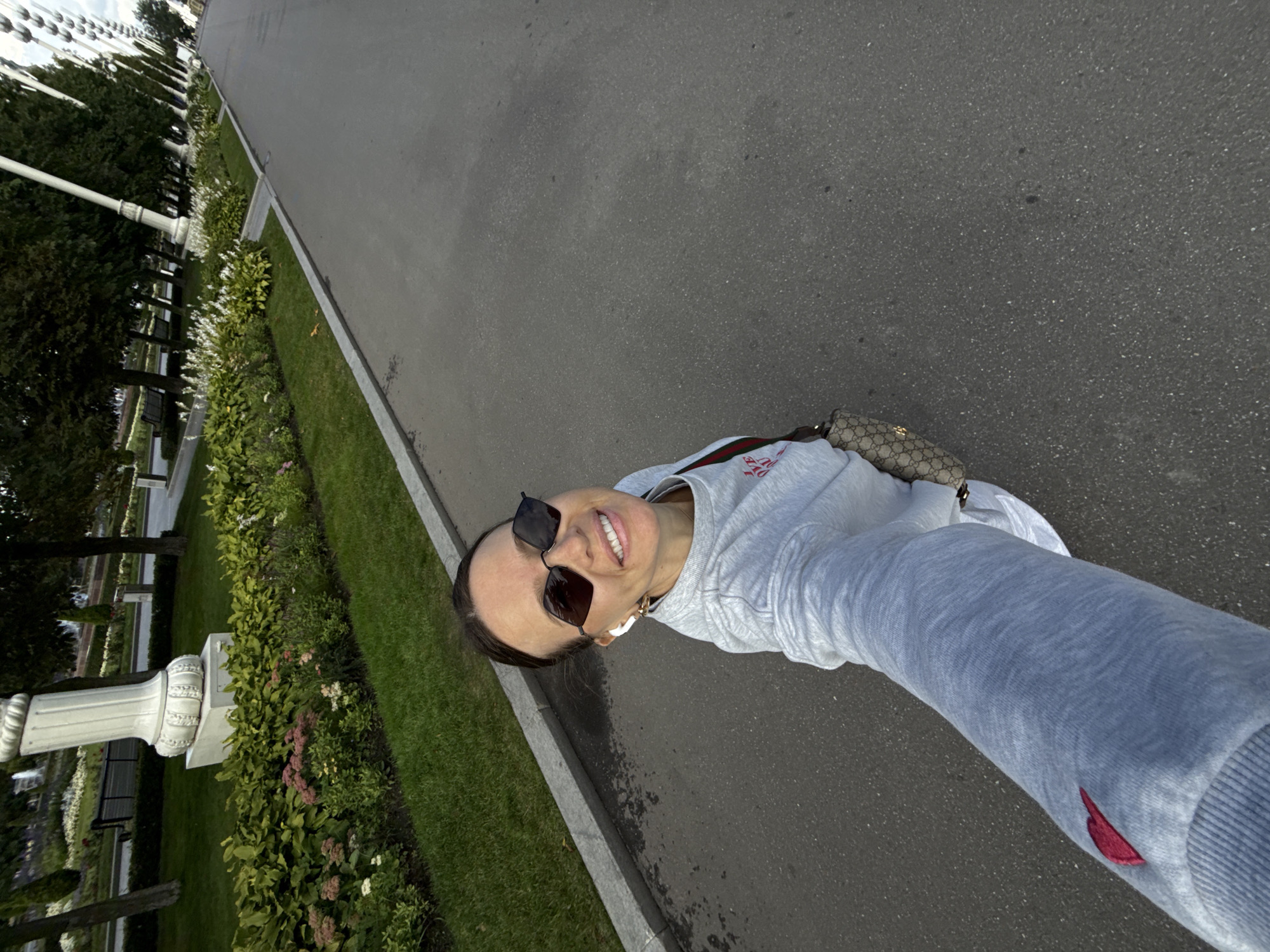 A woman in sunglasses and a gray sweatshirt walks on a paved path in a park. She is taking a selfie with her arm extended. In the background, there are trees, manicured bushes, and decorative pillars. The scene appears to be in the city of Khimki, Russia.