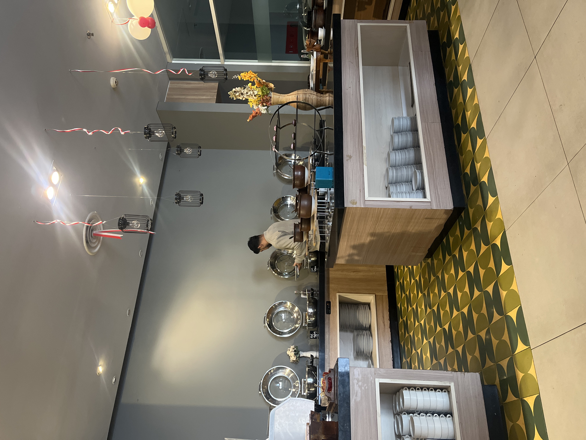 The image depicts a buffet station at a restaurant in Gapurosukolilo, Indonesia. A man in a light-colored shirt is standing behind a buffet counter, attending to food. The buffet features several large, silver chafing dishes with glass lids, likely containing hot food. In front of the chafing dishes, there are stacks of plates and various serving utensils. To the right, a vase with colorful artificial flowers adds a decorative touch. The counter itself is dark, possibly granite, and the cabinets below are made of light-colored wood, displaying stacks of white plates. The floor is tiled with a distinctive pattern of green and yellow geometric shapes. The ceiling has several hanging light fixtures and decorative red and white ribbons, suggesting a festive occasion or theme. The overall environment appears to be an indoor dining area.