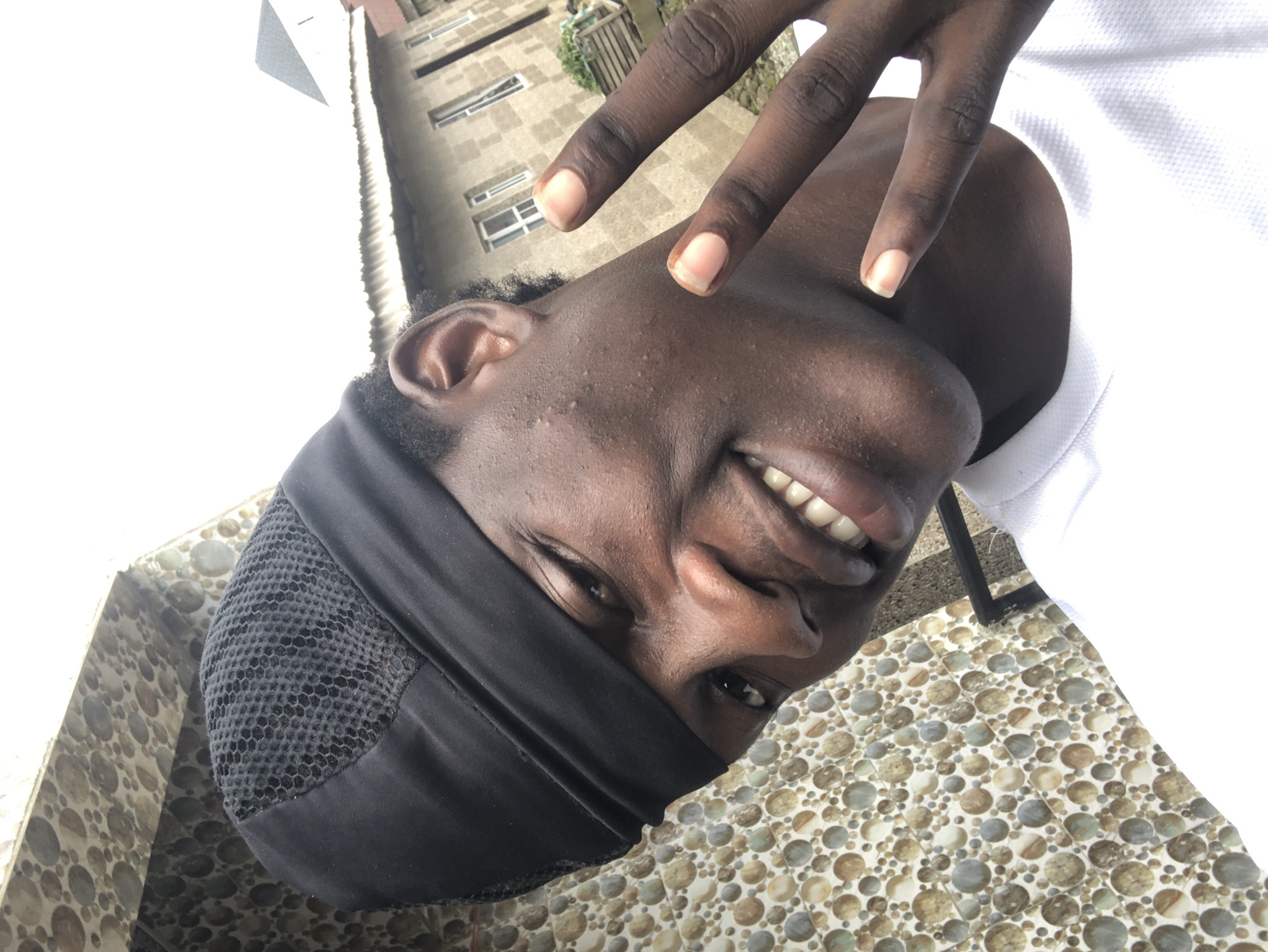 A close-up selfie shows a young man with dark skin and a black durag on his head. He is smiling and making a peace sign with his right hand. He is wearing a white t-shirt. The background consists of a patterned tiled wall and a building with windows. The location is in Ado, Nigeria.