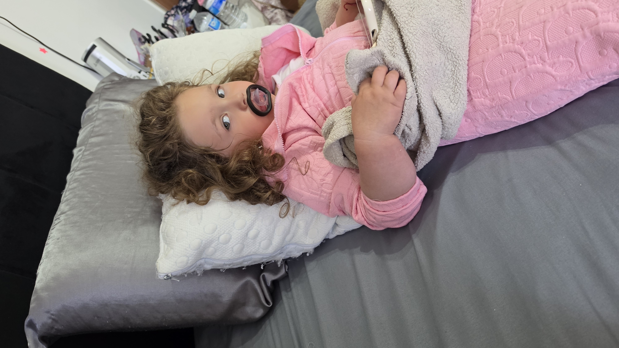 A toddler with curly brown hair lies on a grey bed. The child wears a pink zip-up jacket and has a black pacifier in their mouth. Their head rests on a white pillow with a textured surface. A grey towel is draped over the toddler's body and held in their hand. The bed has a dark headboard visible on the left side. A water bottle and other items are in the background, slightly out of focus. There is no indication in the image that the scene is located in London.