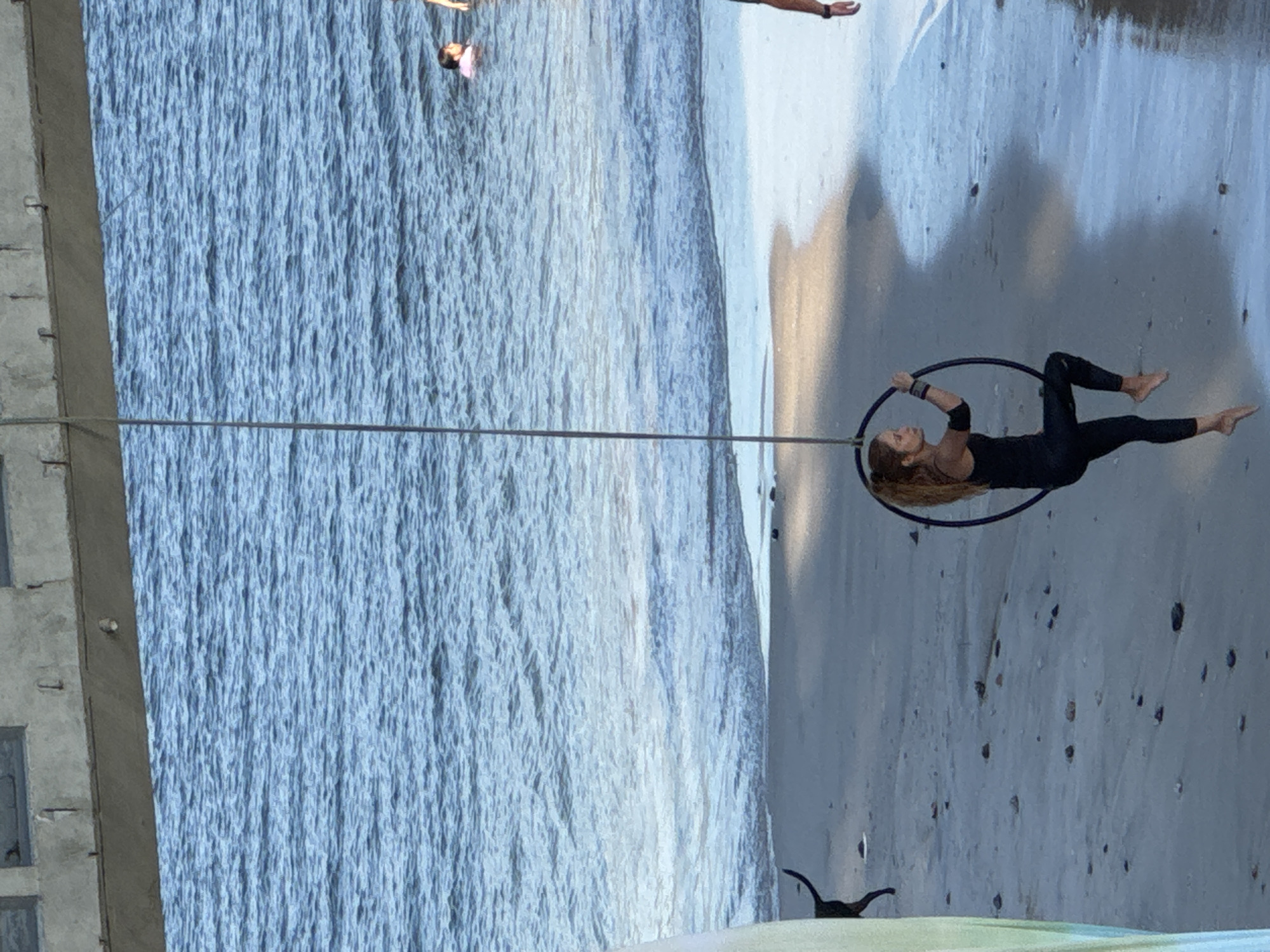 An aerialist performs on a hoop suspended by a rope over a sandy beach in Niterói, Brazil, with the ocean in the background. The aerialist, a woman with long blonde hair, wears a black athletic outfit and wristbands. She is positioned inside the hoop, with one leg bent and the other extended. The hoop is dark colored and appears to be made of metal. The beach is sandy with some scattered pebbles. The ocean is calm with gentle waves lapping the shore. In the distance, a few people are visible in the water. Above the aerialist and the ocean is a concrete structure that appears to be part of a bridge or pier. A thin rope connects the hoop to the elevated structure. A dark-colored dog with its tail raised is visible in the sand to the left of the aerialist.