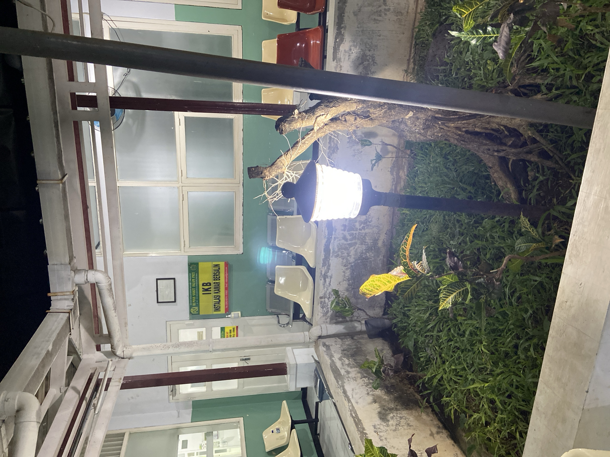 This media file depicts an outdoor waiting area at a facility, possibly a hospital or clinic, in Margoyoso, Indonesia. The scene is lit by a warm-toned outdoor lamp, suggesting it is either dusk or nighttime. Greenery, including grass and potted plants with variegated leaves, surrounds the waiting area.
The waiting area itself features several rows of seating. Some chairs are simple, molded plastic in a light beige color, while others are darker, possibly red or maroon, and appear to be made of metal and plastic. The architecture suggests an open-air or semi-enclosed space with a roof supported by white pillars and metal beams. Visible pipes run along the exterior wall.
A sign in Indonesian is prominently displayed on the green wall. It reads: "RUMAH SAKIT ISLAM PATI" at the top, followed by "IKB INSTALASI KAMAR BERSALIN". Below this, there is smaller text with a phone number and email address. Another smaller yellow sign is partially visible with the word "MAAF".
The overall impression is one of a functional waiting space designed to be illuminated and accessible during darker hours, with a touch of natural landscaping. The presence of the sign for "INSTALASI KAMAR BERSALIN" (Delivery Room Installation) strongly suggests a medical facility, specifically a maternity ward. The lighting creates a somewhat subdued and peaceful atmosphere.