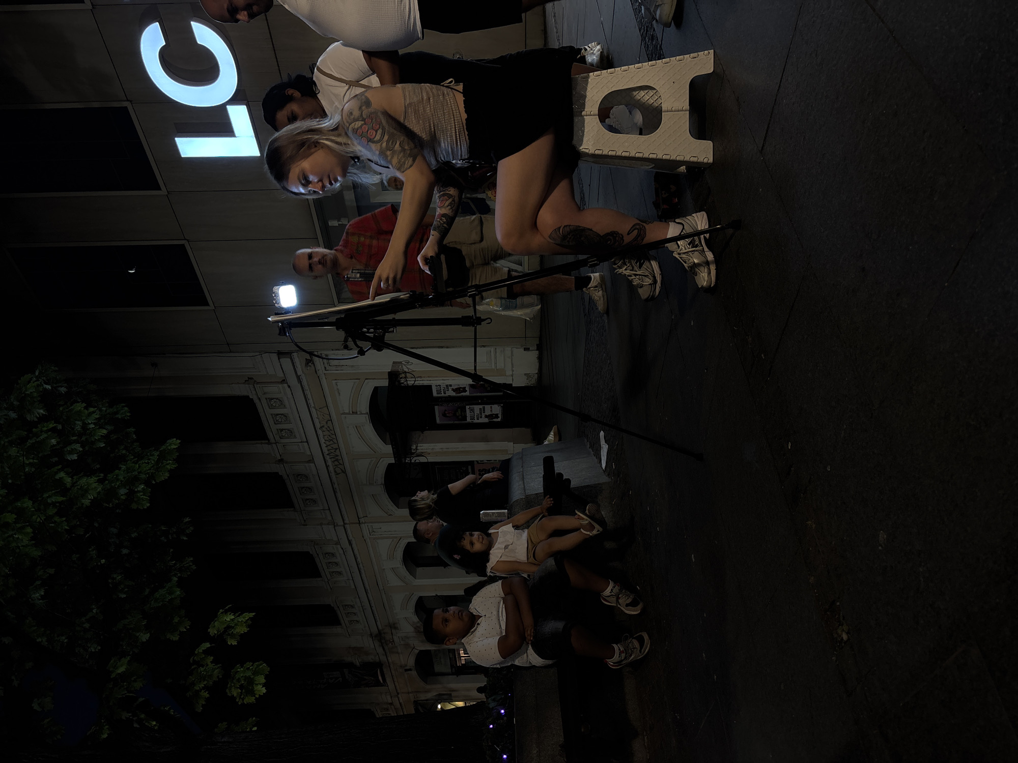 The image depicts a night scene in Belgrade, Serbia, likely on a pedestrianized street or a plaza. The background features a building with arched windows and signage reading "LC" in large, illuminated letters. In the foreground, a young woman is seated on a portable stool, focused on a drawing or painting on an easel. She has numerous tattoos on her arms and legs. A man in a red shirt and a woman in a white shirt stand behind her. To the left, two children, a boy and a girl, are seated on a stone bench. The boy is wearing a white polo shirt and denim shorts, while the girl is in a white top and light-colored shorts. The overall atmosphere is relaxed, with a street artist engaging with her work and a small audience or passersby observing. The lighting is dim, with artificial lights from the building and a spotlight on the artist's easel creating illumination.