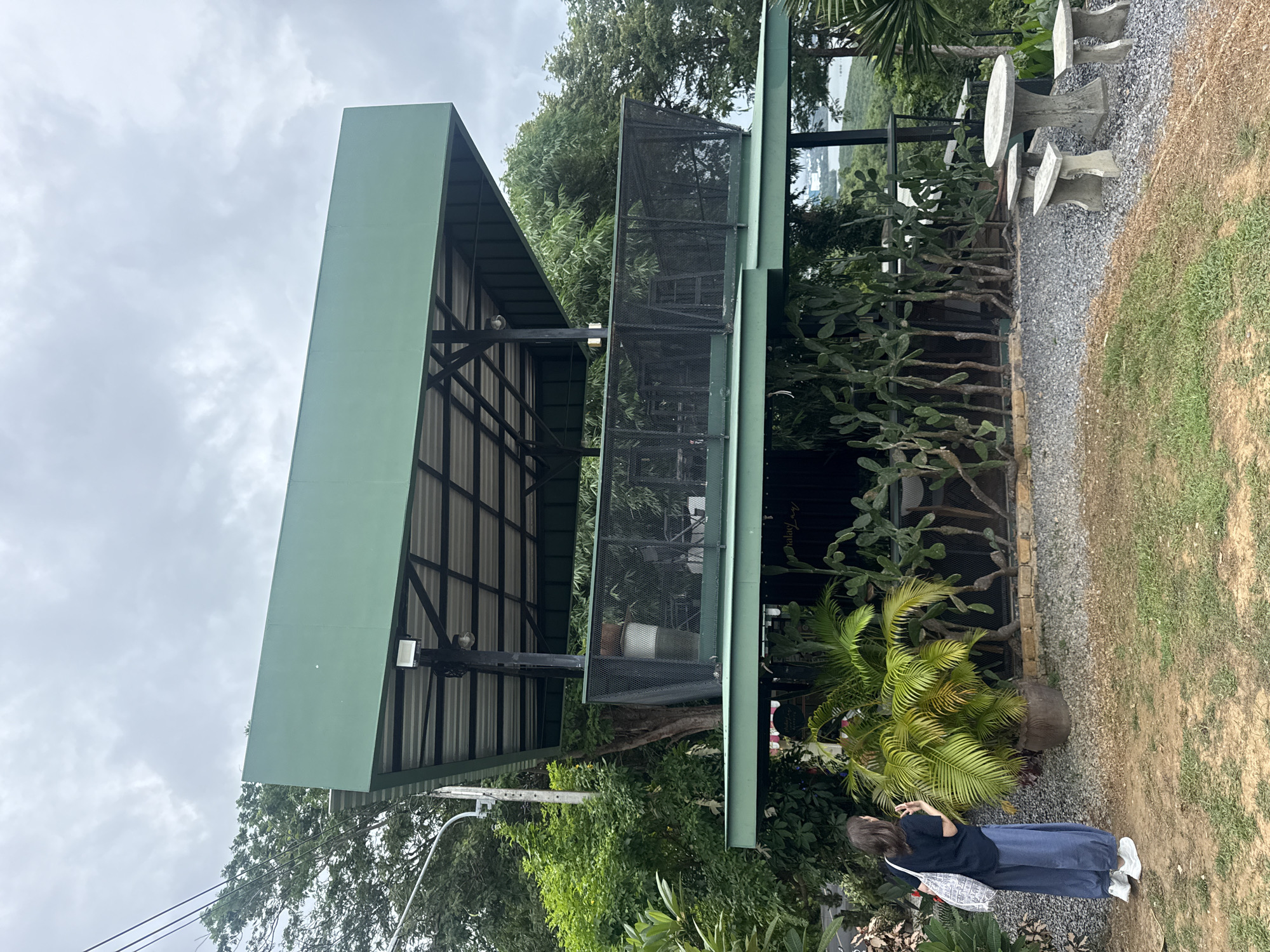 A two-tiered green building with a corrugated metal roof stands on a slight incline, surrounded by lush vegetation. The lower level features a wall lined with a variety of cacti and potted plants, with a sign visible on the wall. A person wearing a dark blue shirt and loose blue pants stands to the left of the building, facing away from the camera, holding a patterned tote bag. In front of the building, a concrete table and four accompanying stools are arranged on a gravel surface. In the background, trees and a body of water are visible under a cloudy sky. The location is identified as Wichit, Thailand.