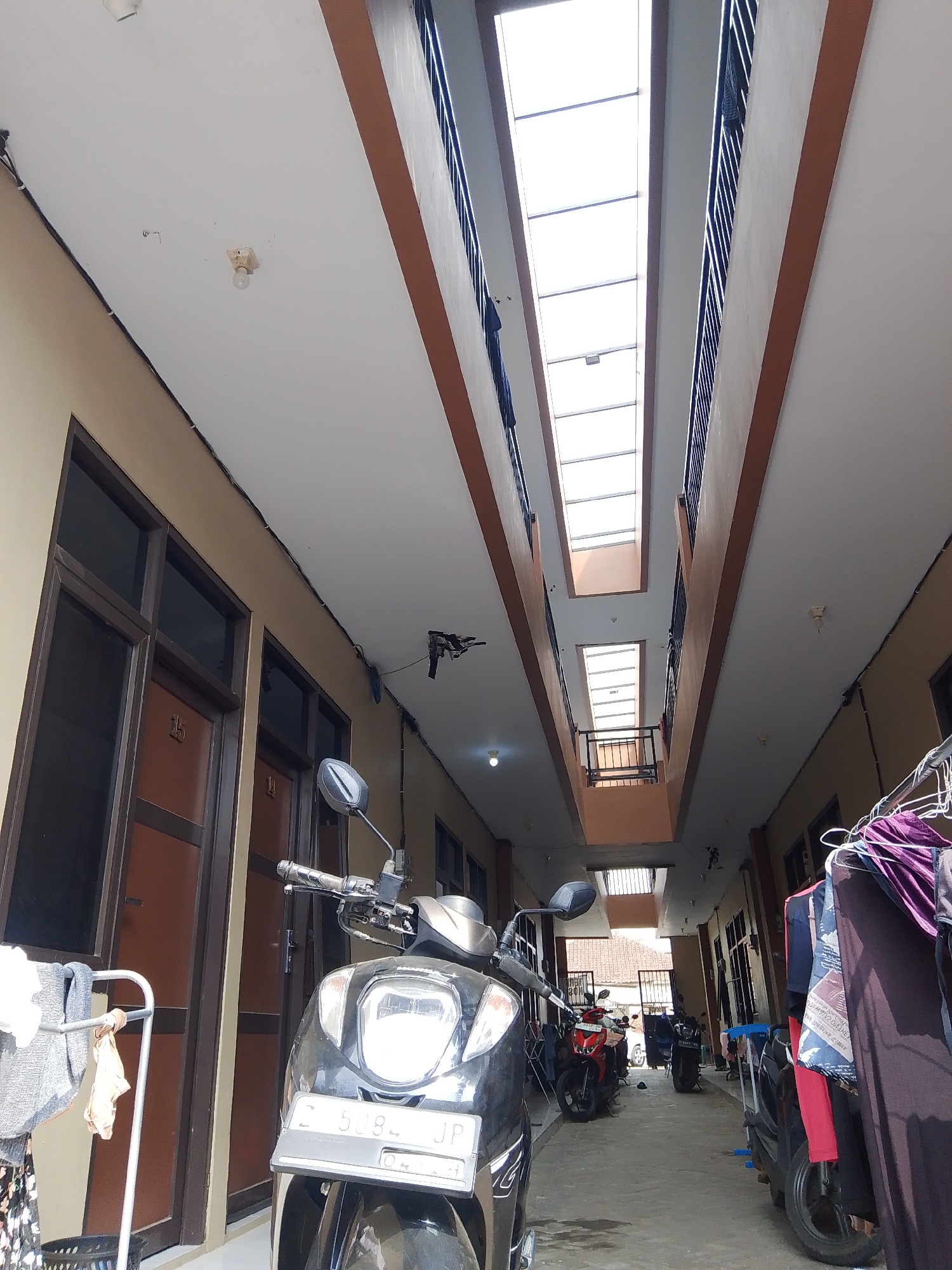 The image depicts a narrow, open-air corridor between two multi-story buildings. The corridor is covered by a translucent roof with skylights, allowing natural light to enter. The buildings have multiple doors, some with visible room numbers like "15" and "14." Several motorcycles are parked along the corridor. On the right side, laundry is hanging on a rack, with various garments displayed. The environment appears to be an urban setting, likely a residential complex or a dormitory, given the row of rooms and parked vehicles. The architecture suggests it could be located in Tasikmalaya, Indonesia, a city known for its densely populated areas and apartment-style housing. There are no people visible in the frame.