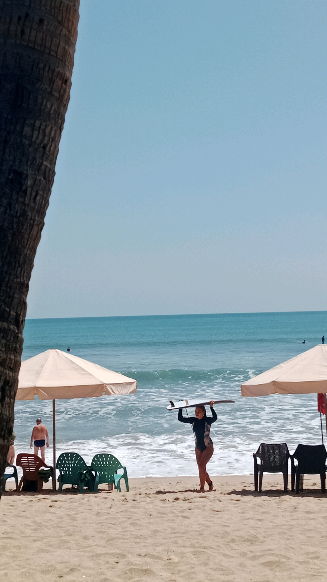 A wide-angle, vertical shot captures a beach scene in Kuta, Indonesia. In the foreground, the textured trunk of a palm tree dominates the left side of the frame. The sandy beach stretches across the bottom, featuring several beach chairs and two large, cream-colored umbrellas.
To the left, under an umbrella, a man in blue swimming trunks stands facing away from the camera. In the center, three green plastic chairs are arranged, with a reddish-brown chair to their left.
Further into the scene, a woman in a black wetsuit is walking across the sand, carrying a surfboard over her head. She is facing the right side of the frame, her body angled slightly towards the viewer. Her wetsuit features a patterned design on the leg.
In the background, the turquoise ocean meets the horizon under a clear, light blue sky. Waves are breaking on the shore, creating white foam. A few other figures are visible in the water and on the beach in the distance. The overall atmosphere is one of a sunny day at a tropical beach resort.