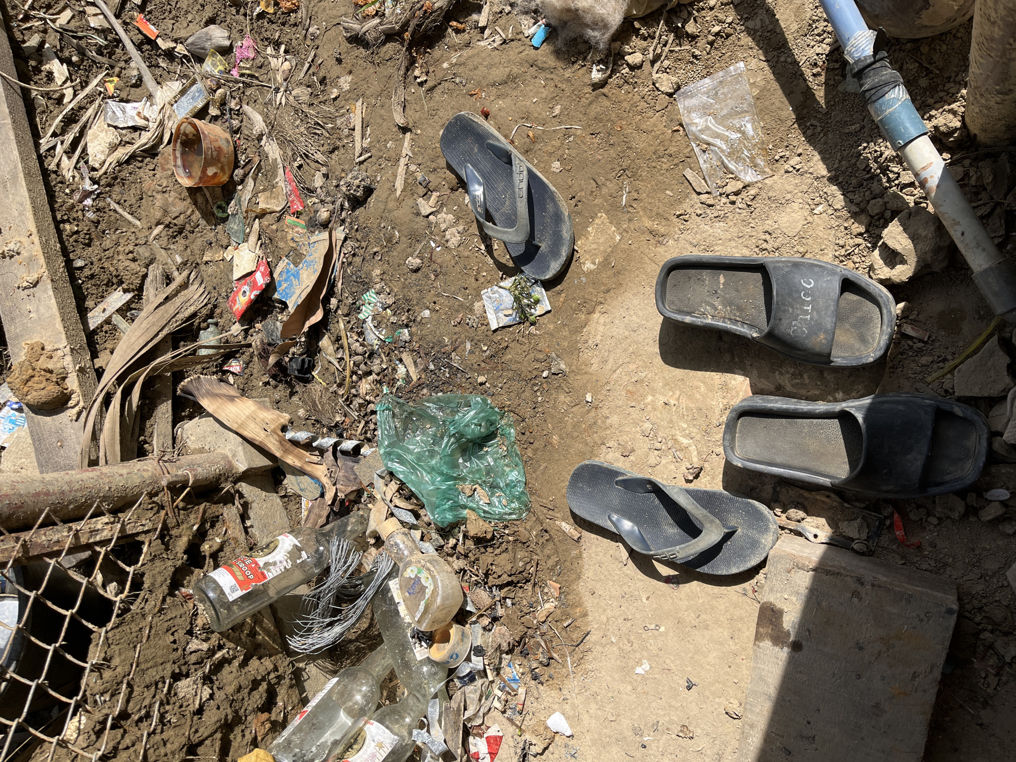 The image depicts a cluttered, outdoor scene on uneven, muddy ground, possibly in a less maintained area of Kota Kuala Simpang, Indonesia. Scattered debris includes broken glass bottles, plastic bags, torn packaging, dried leaves, and what appears to be wire. In the foreground and midground, three pairs of dark-colored footwear are visible: two pairs of flip-flops and one pair of slides. One pair of flip-flops is positioned to the left, with the sole facing upwards. Another pair of flip-flops is situated in the mid-right of the image, lying flat. A pair of black slides, with the word "Chicc" embossed on the strap, rests in the lower right quadrant. A rusty, partially submerged pipe runs across the bottom right of the frame. The lighting suggests a sunny day, casting distinct shadows. The overall impression is one of neglect and accumulation of waste.