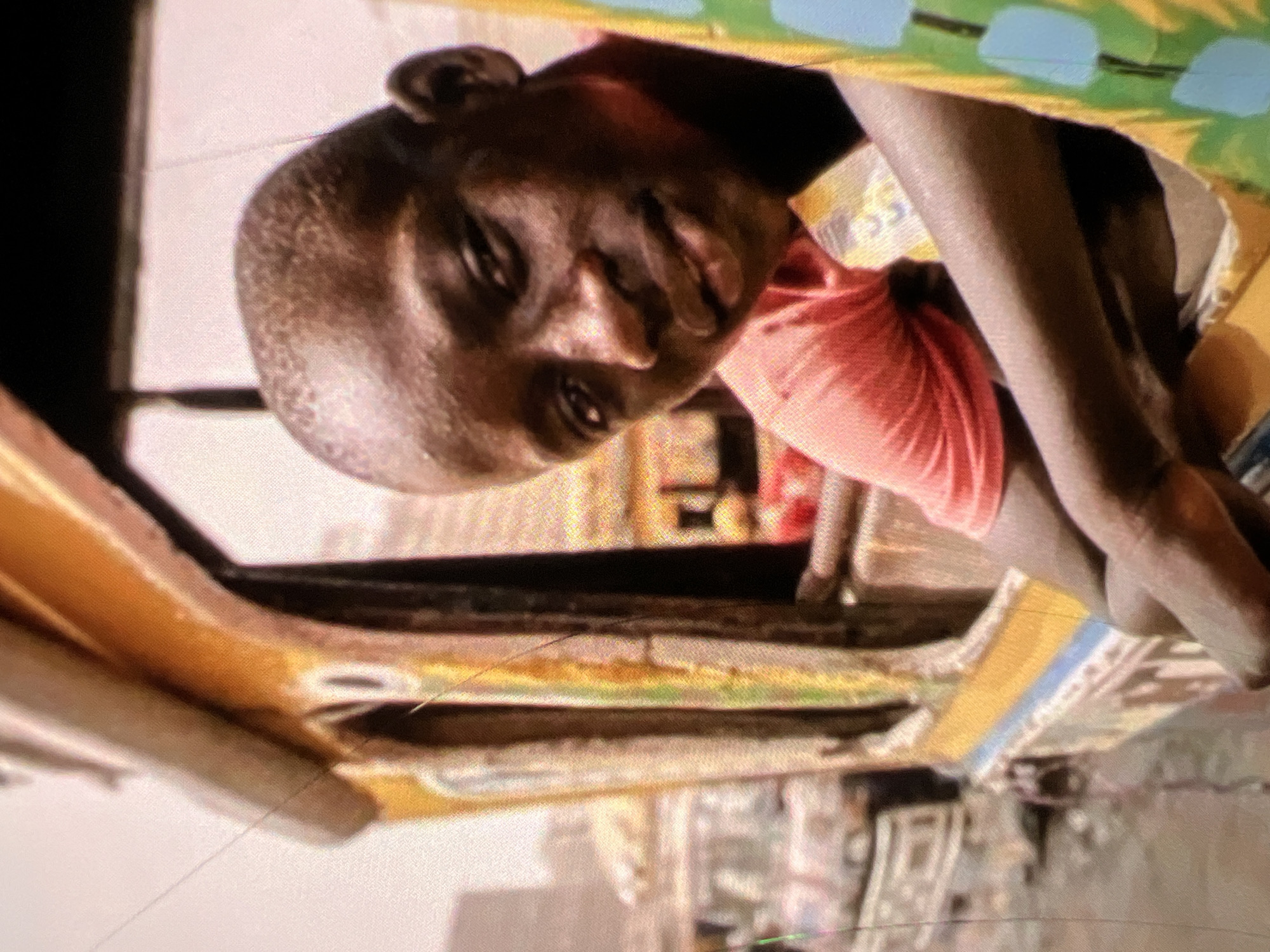 The image shows a close-up of a young Black boy leaning out of a window of a brightly painted bus. The bus appears to be in motion, suggesting an urban environment. The boy is wearing a salmon-colored t-shirt and is looking directly at the camera with a neutral expression. His arms are resting on the edge of the window frame, and his hands are visible. The interior of the bus is visible in the background, along with other elements of the bus's exterior, which are decorated with various colors and patterns. The background is blurred, but it suggests a bustling city scene with other vehicles and buildings, consistent with an urban setting like Lagos, Nigeria. The image quality is grainy, indicating it may have been taken with a mobile device or in low light conditions.