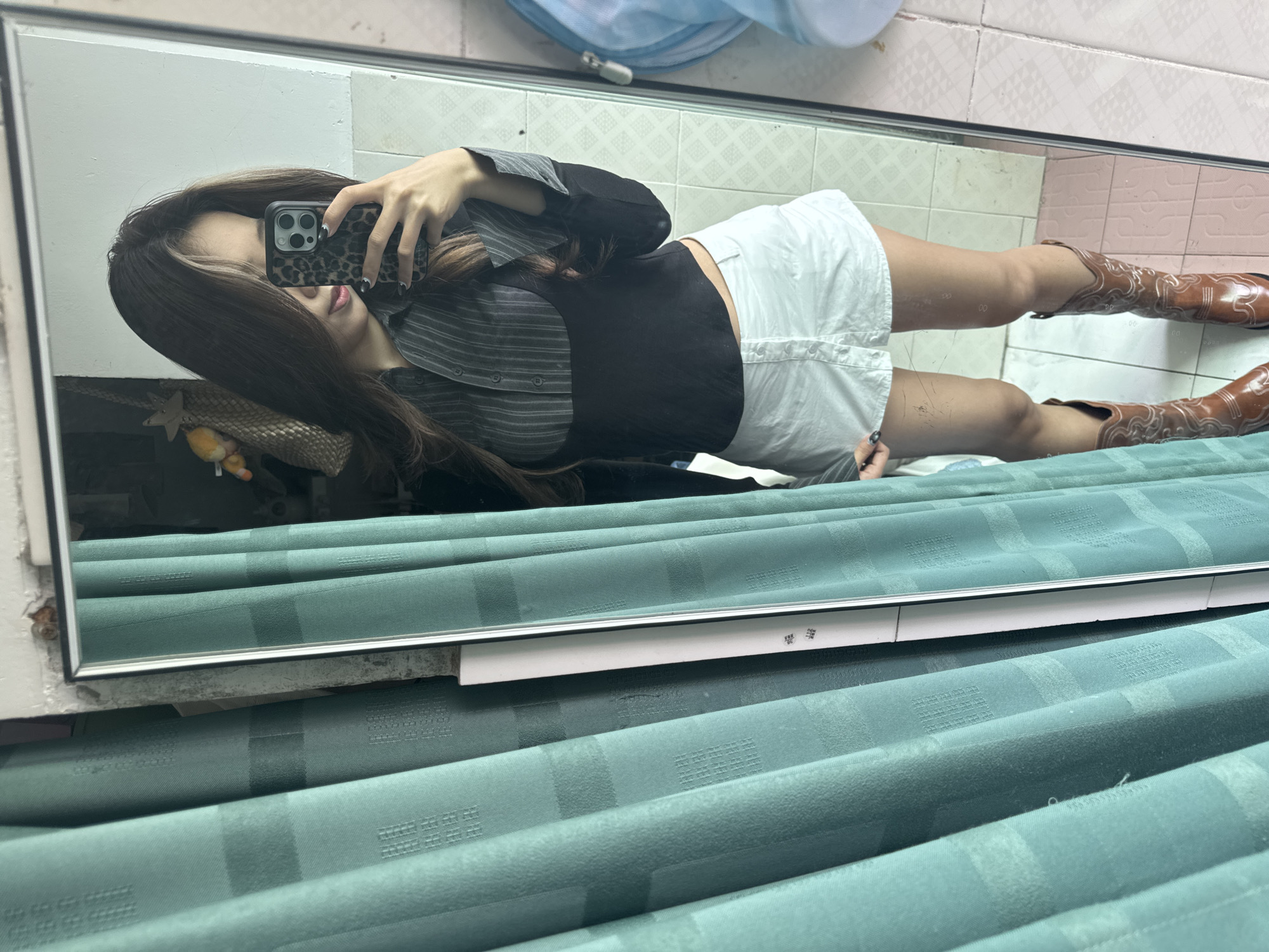 A woman is taking a mirror selfie. She is wearing a dark top, white shorts, and brown boots. She is holding a phone with a leopard print case. The mirror is positioned against a tiled wall. Below the mirror are green patterned curtains. The location is Nan'an District, China.